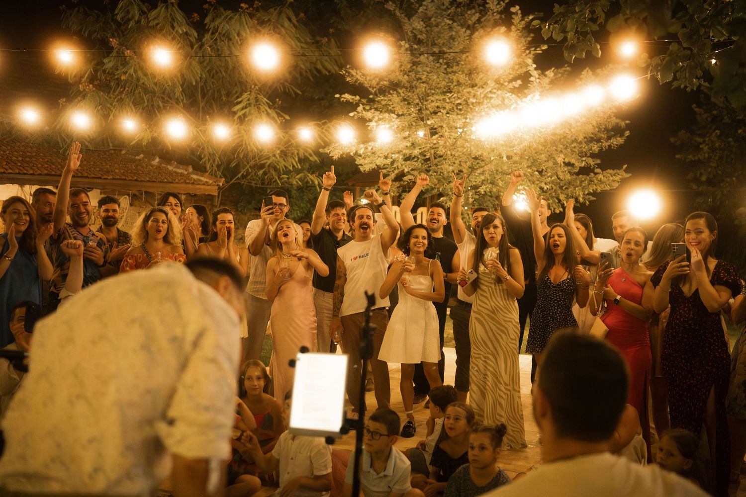 Wedding guests dance under warm string lights at an outdoor evening reception celebration.
