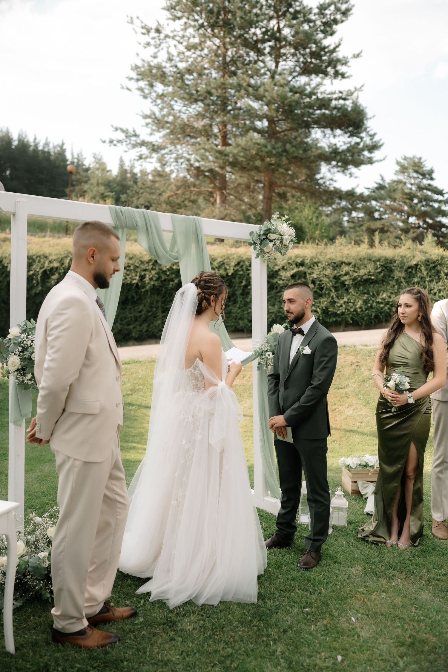 Wedding party gathered outdoors with flowing white curtains and greenery creating an elegant outdoor ceremony setting.