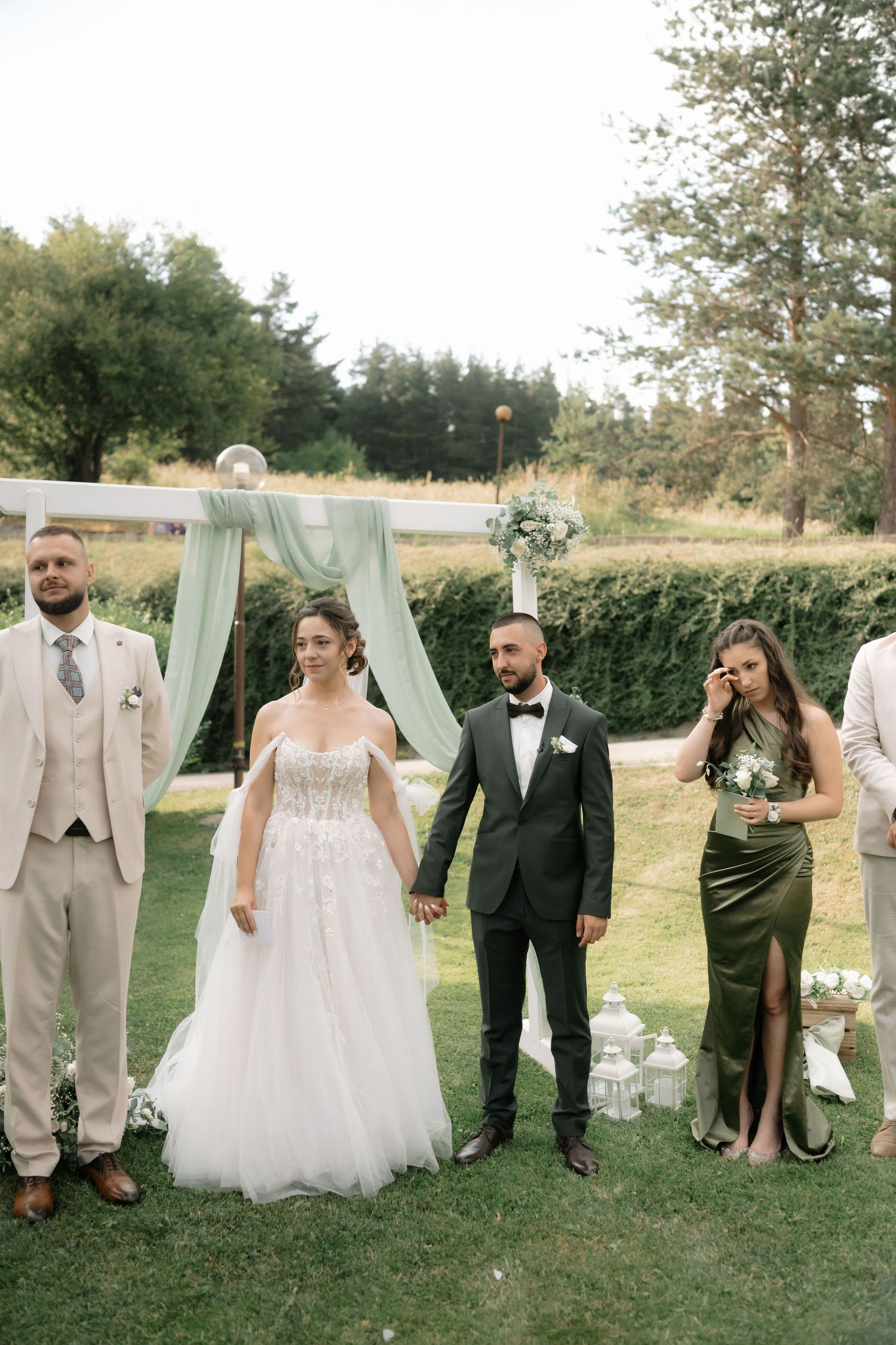 Wedding party members playfully peek through an outdoor draped arch setup in a garden setting.