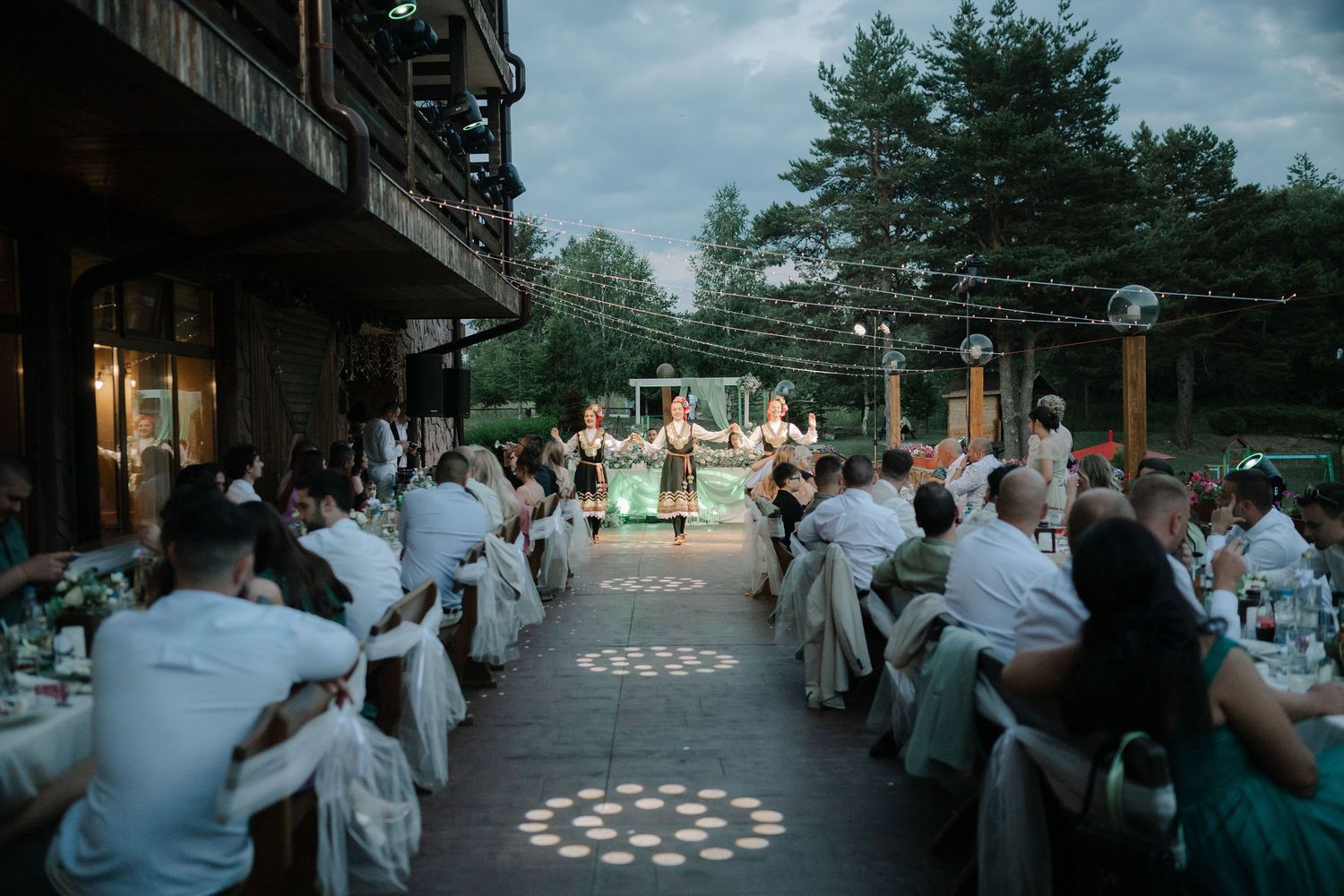 People in white robes gather outdoors at dusk around illuminated circular patterns on the ground during a ceremony.