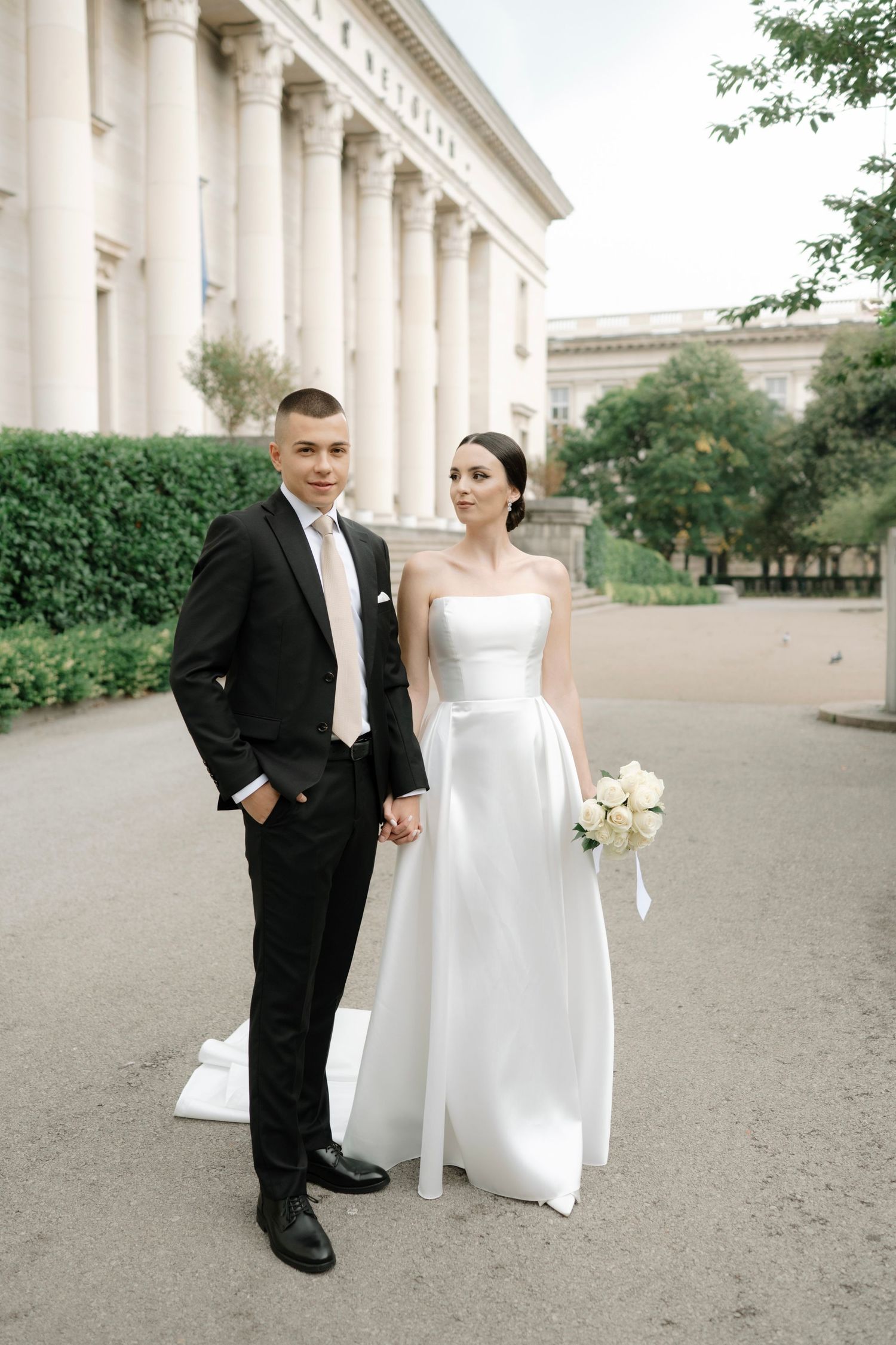 A couple in wedding attire poses in front of a grand building with white columns.