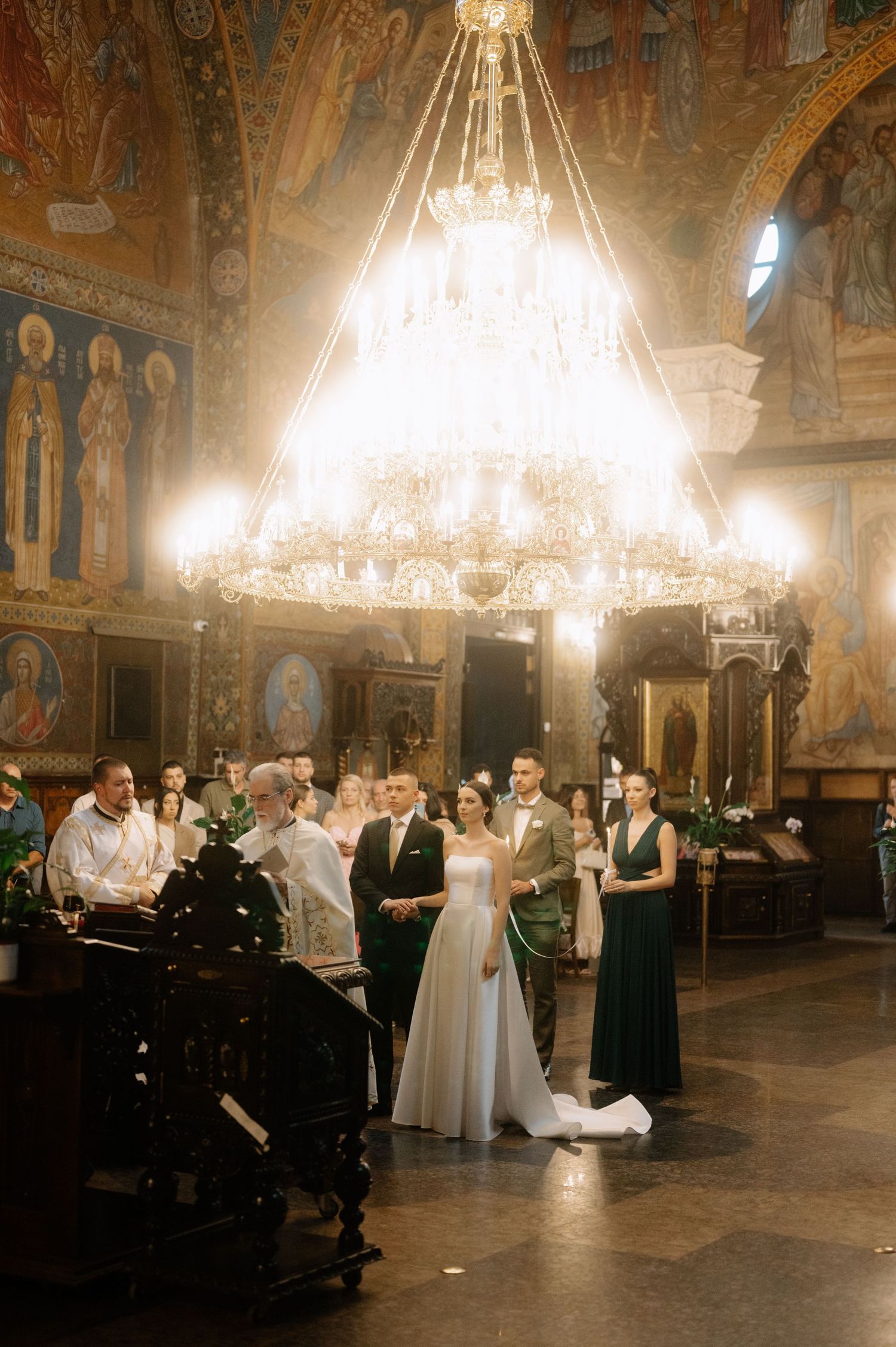 A wedding ceremony takes place in an ornate church hall beneath a grand crystal chandelier with religious artwork on the walls.