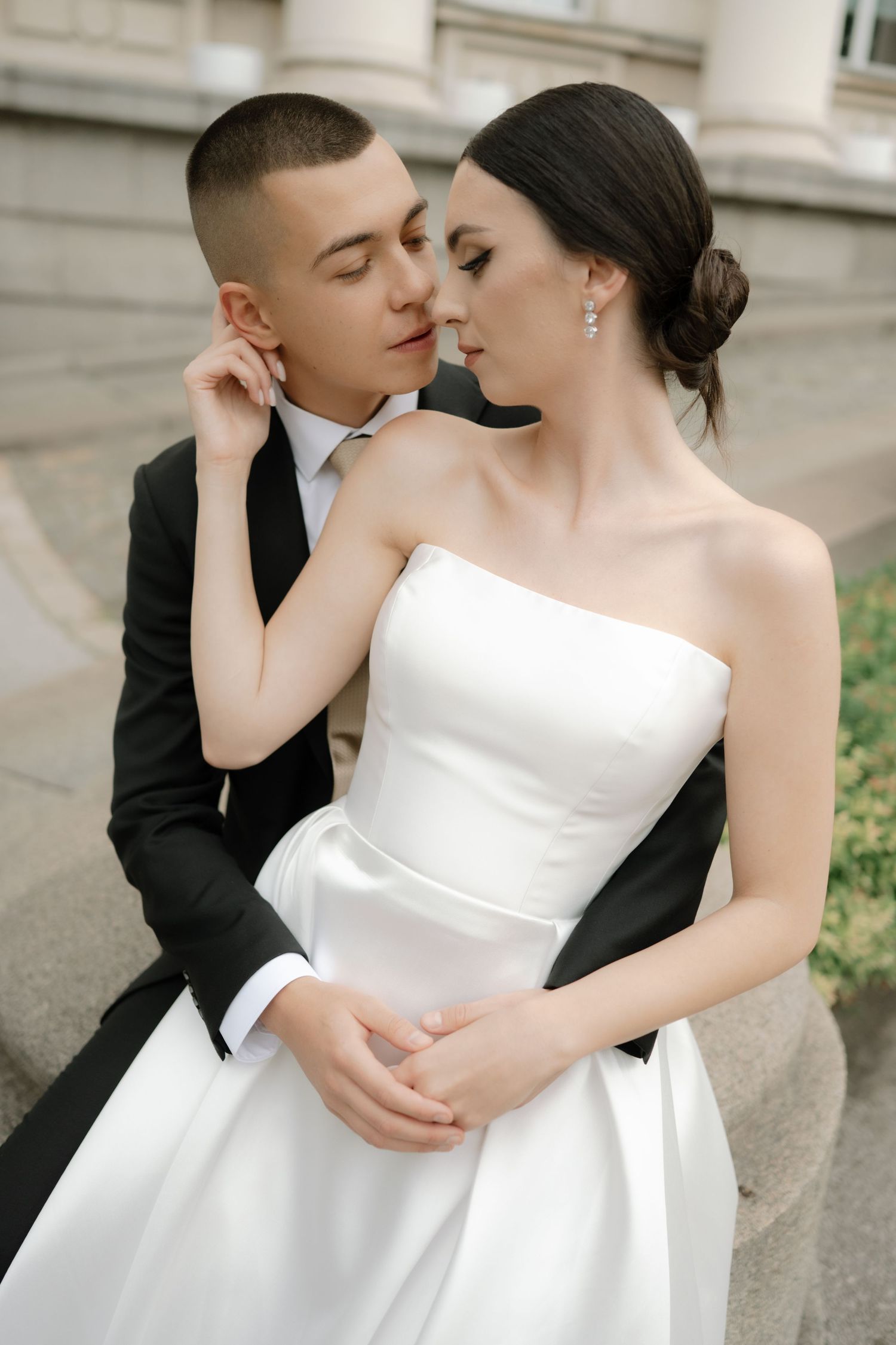 A romantic wedding couple in classic attire share an intimate moment on stone steps, with the bride in a strapless white gown.