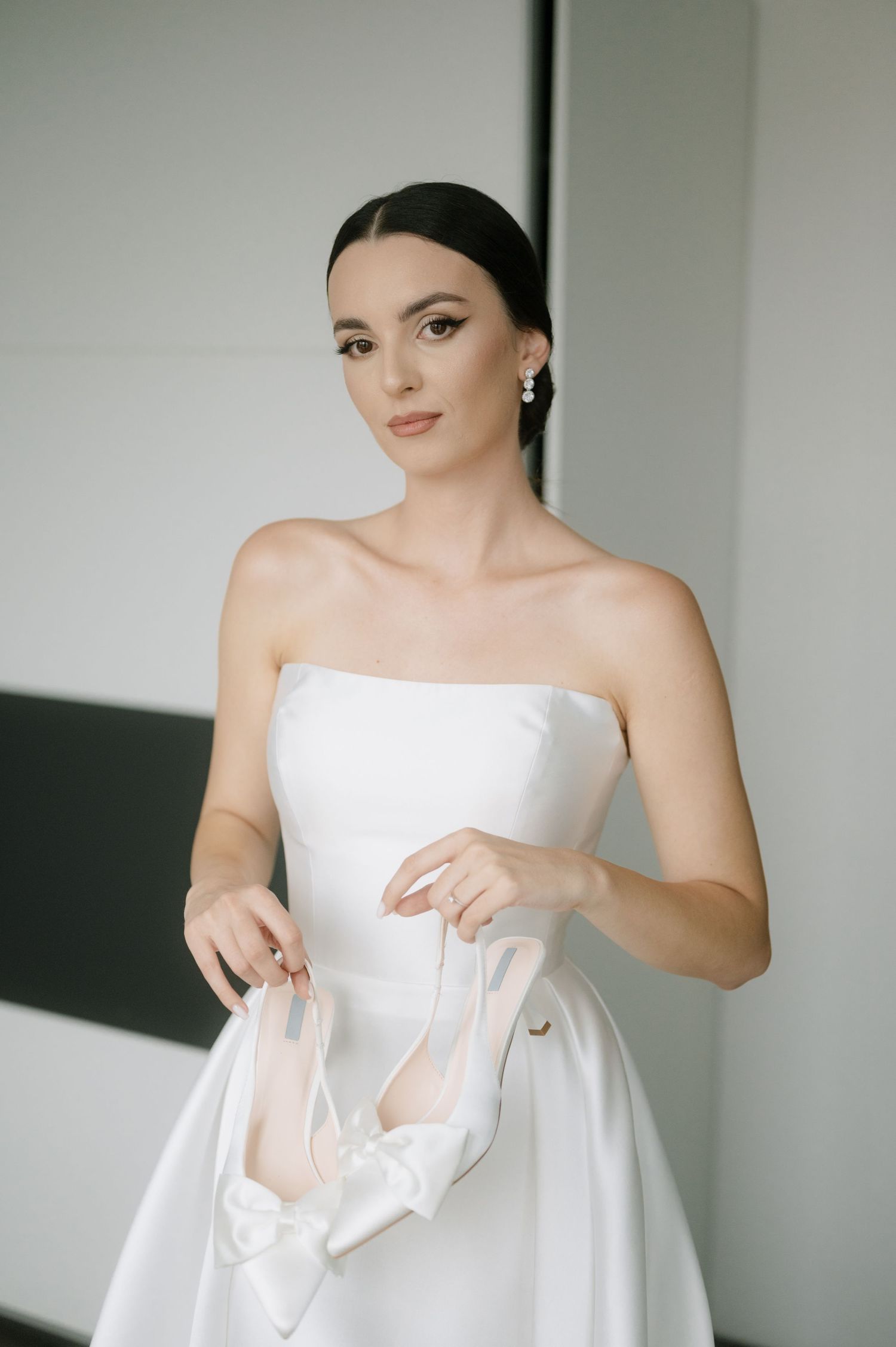 A bride in a strapless white wedding dress adjusts her gown in a minimalist setting.