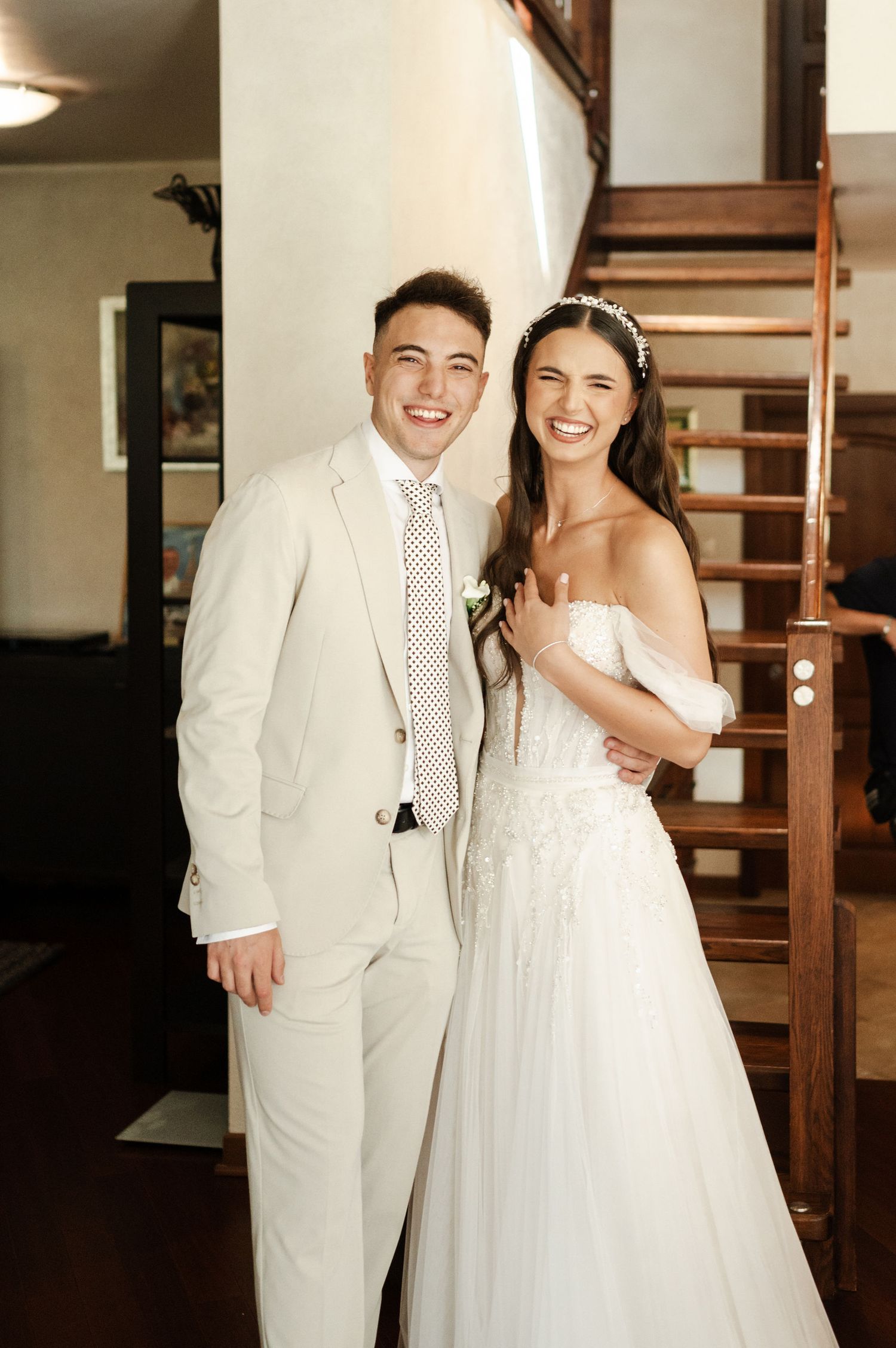 A couple poses for wedding photos in white attire inside a building with wooden stairs.
