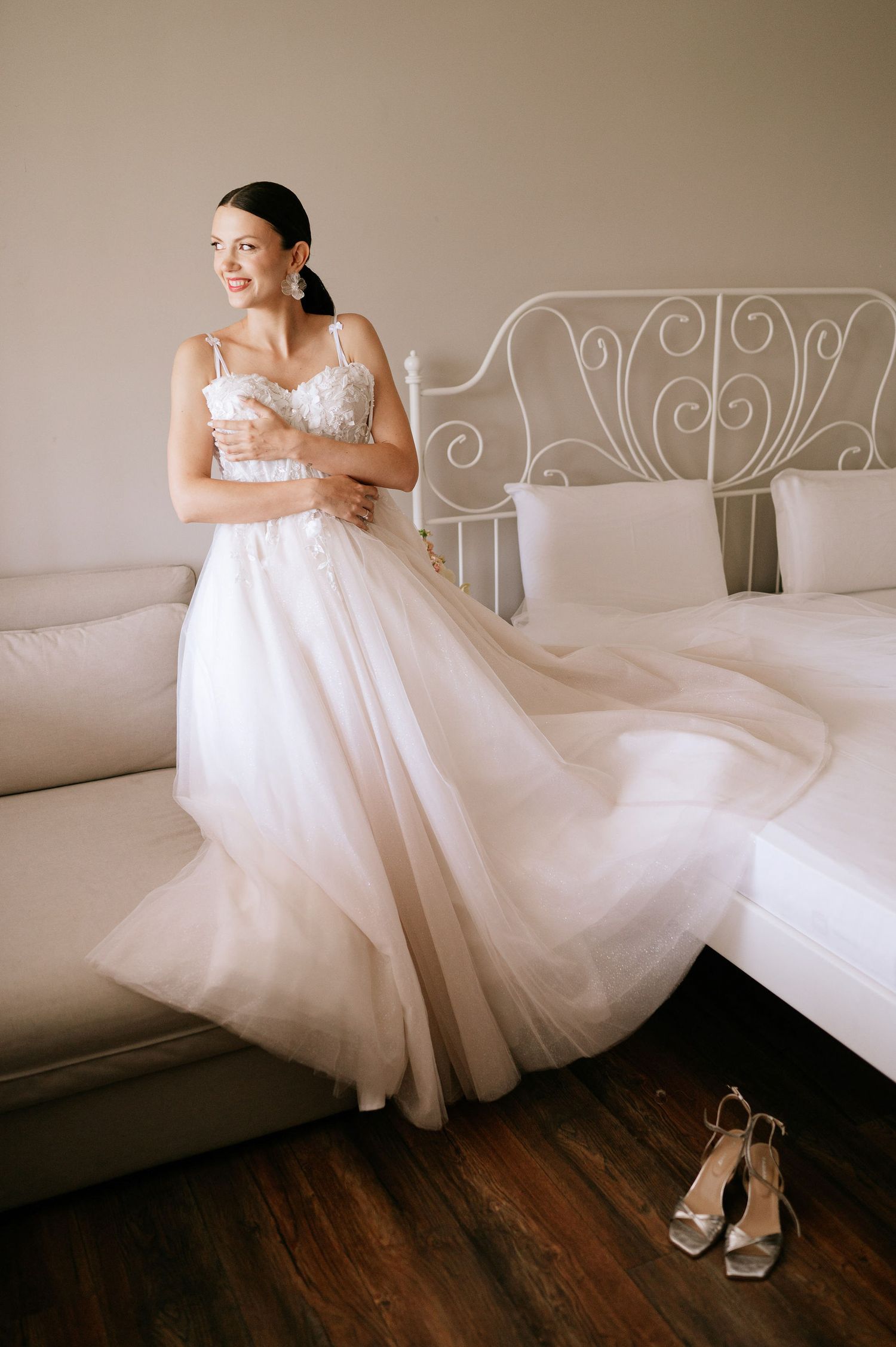 Flowing white wedding gown displayed on an ornate iron bed frame with hardwood floors.