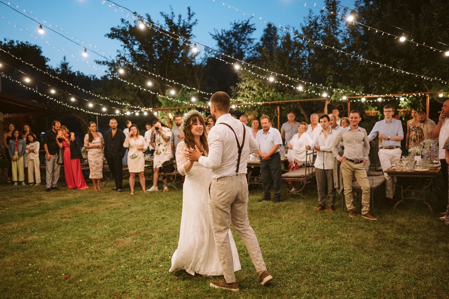 First dance at outdoor wedding reception with string lights and guests watching.