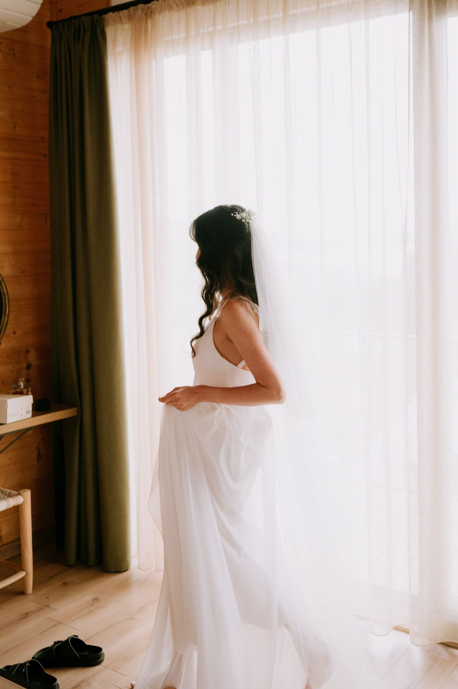 Figure in flowing white dress stands near bright window in wooden room.