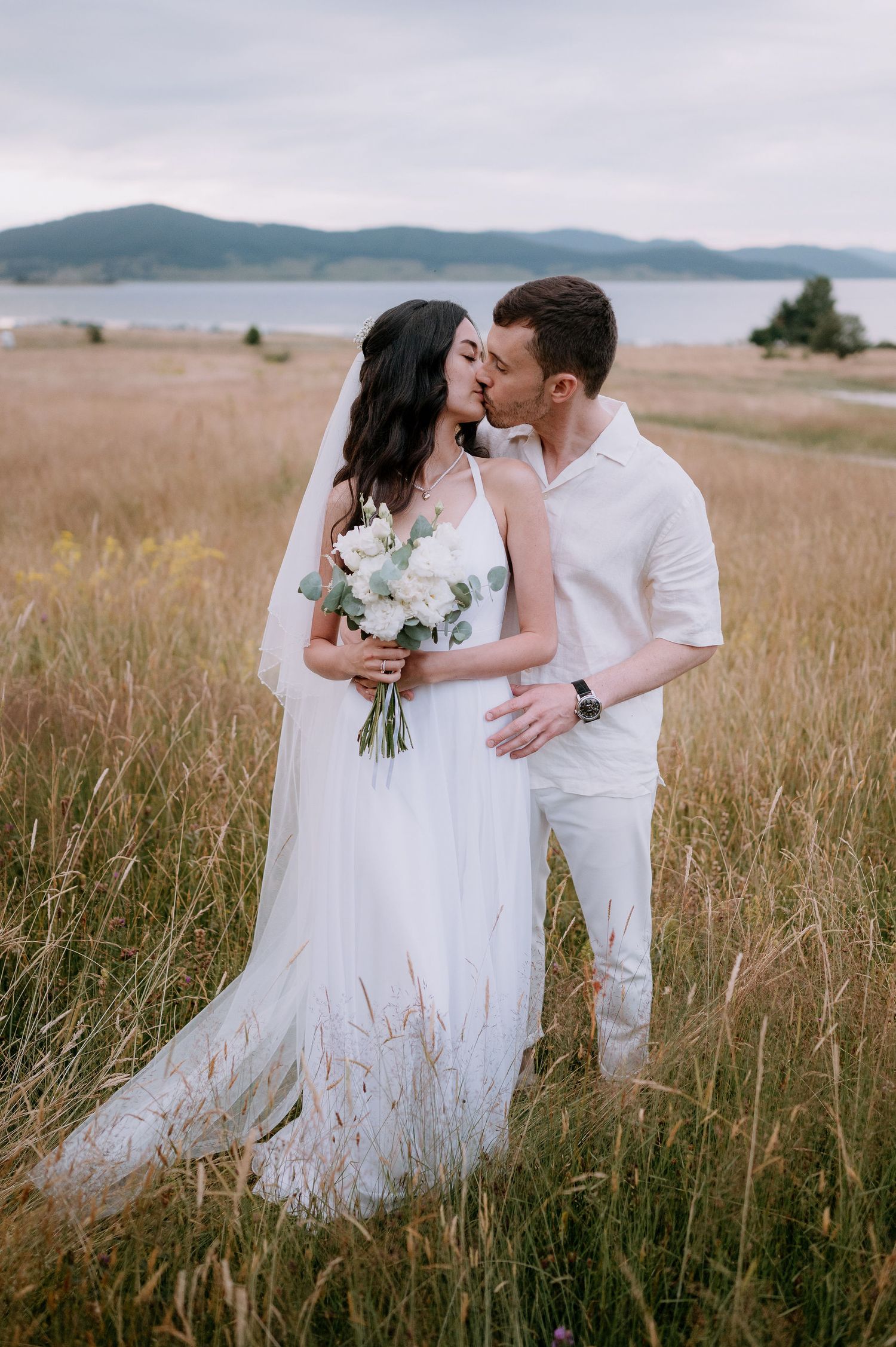 A romantic wedding couple embraces in a golden field with mountains in the background.