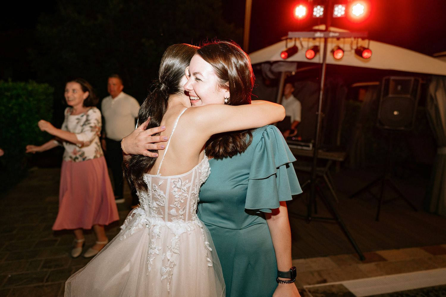 Bride in white dress hugs guest during evening reception celebration.
