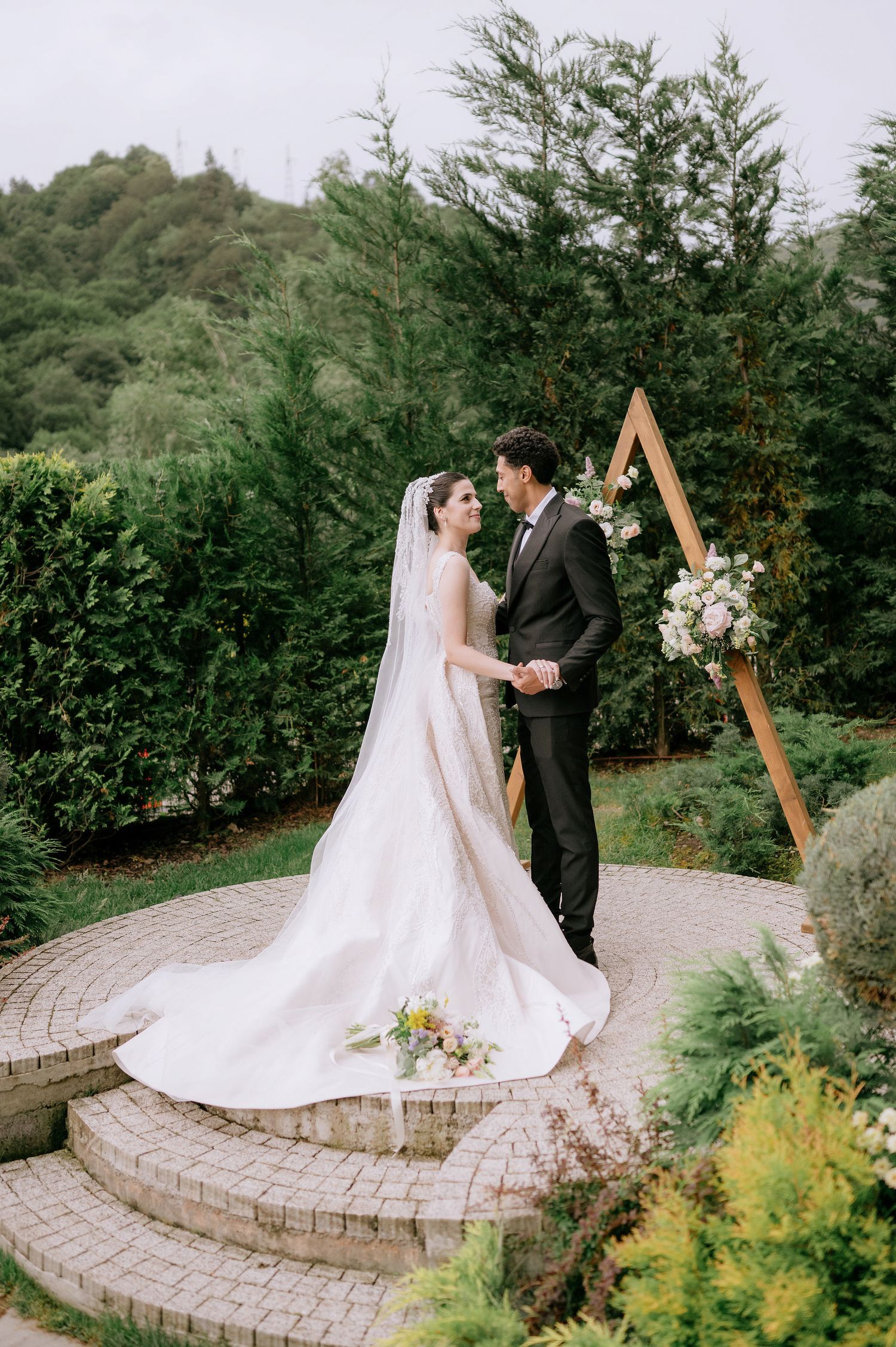 Wedding couple standing on a circular stone patio surrounded by lush greenery and flowers.