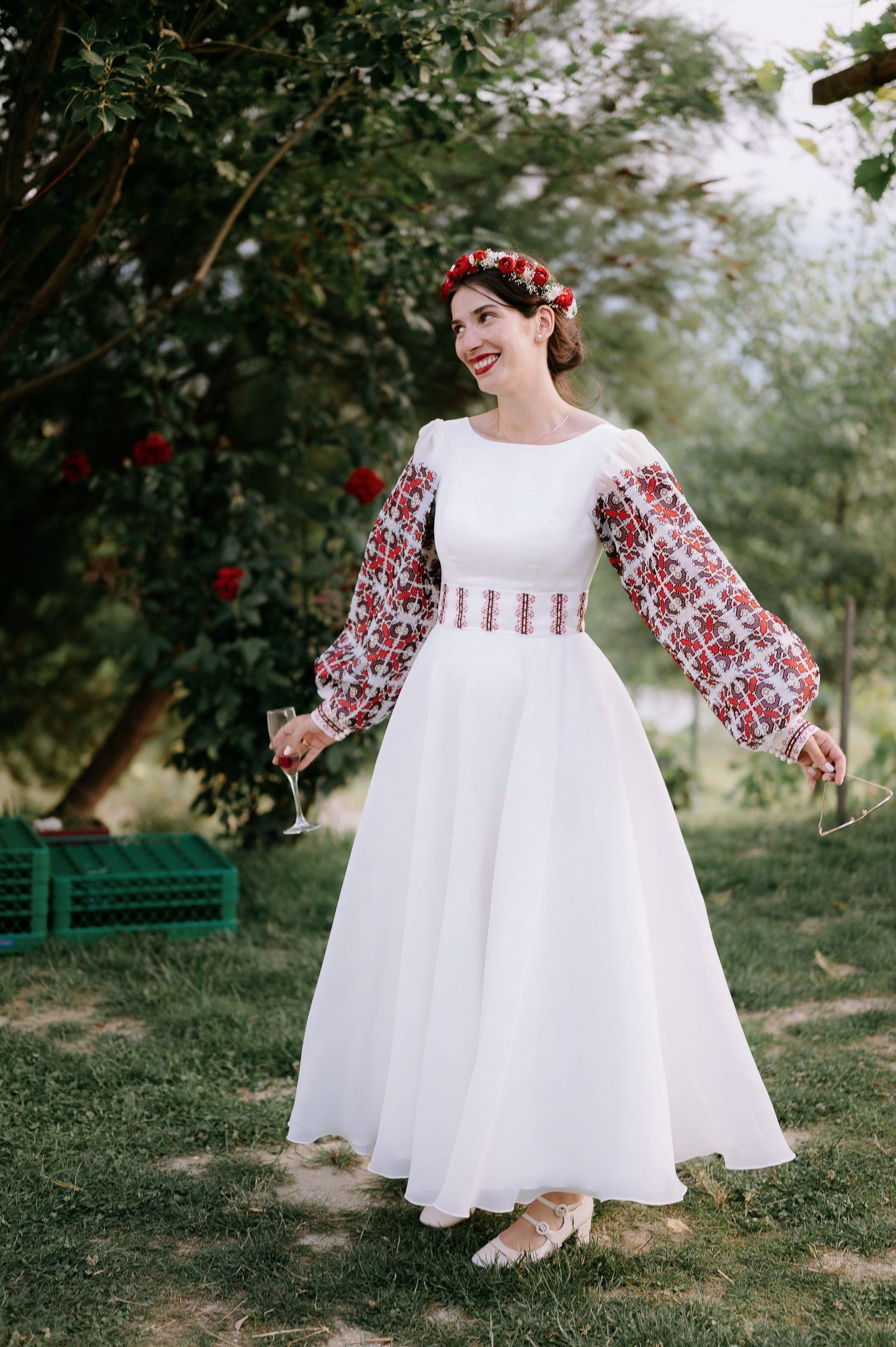 A white dress with traditional red embroidered details flows elegantly in a garden setting.