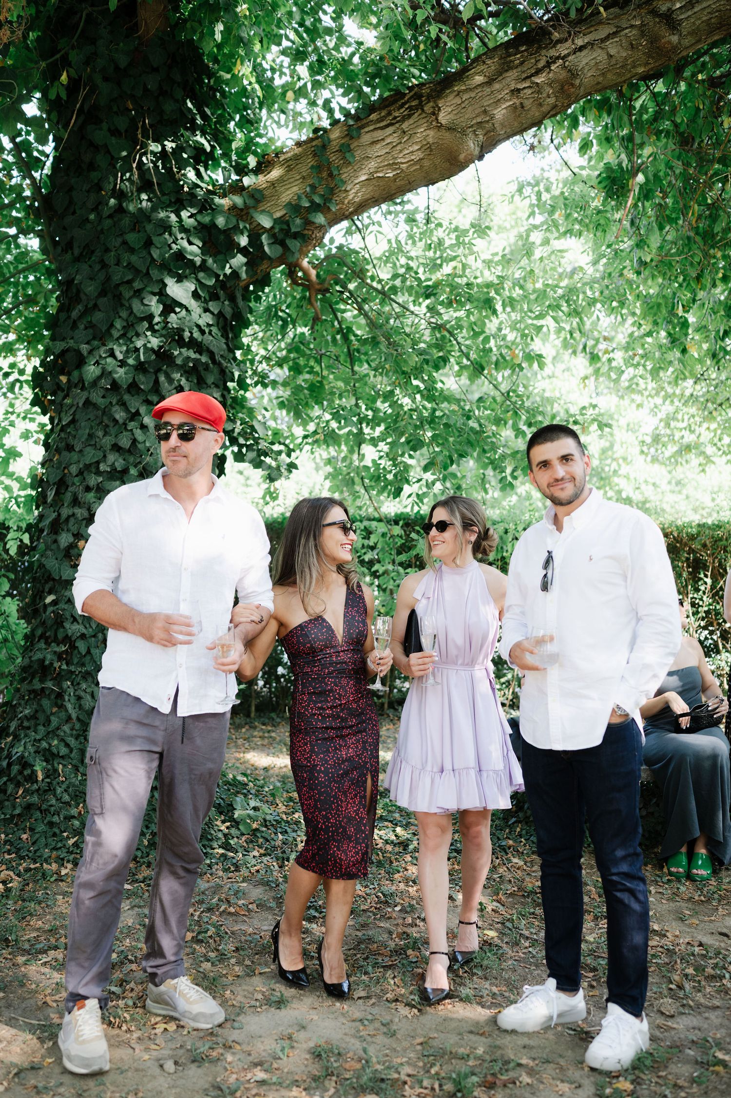 Group of wedding guests gathered under shady trees for casual conversation.