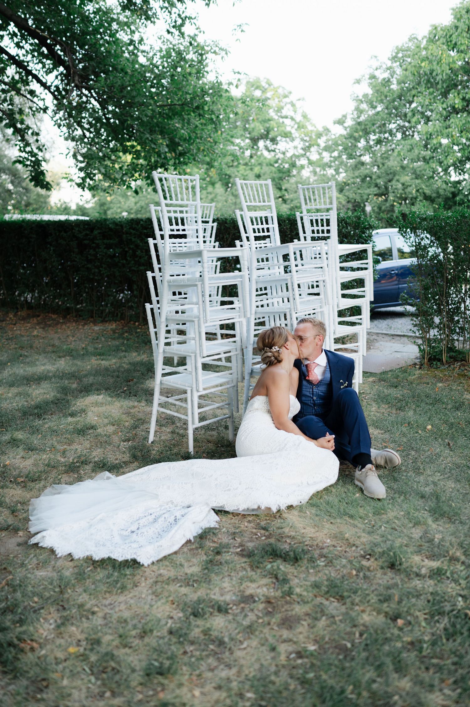 Couple in wedding attire poses casually on grass with white chairs arranged for outdoor ceremony.