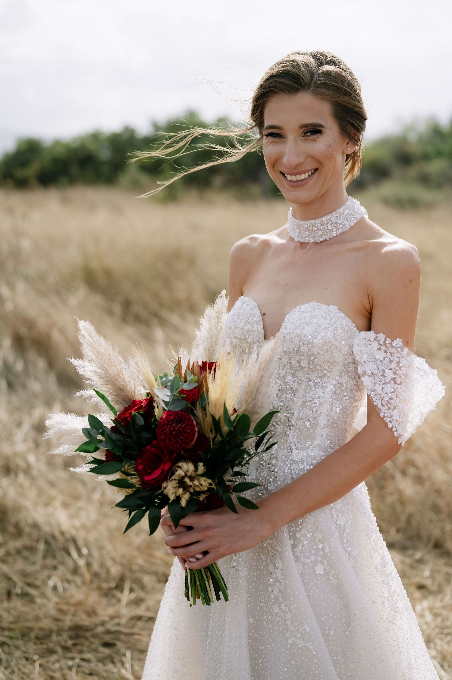 A joyful bridal portrait featuring a white beaded gown and statement burgundy bouquet in a rustic field.