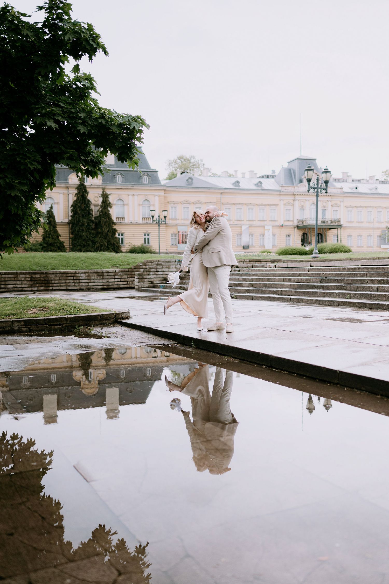 Graceful dancers in neutral tones perform near reflecting pool at historic palace venue.