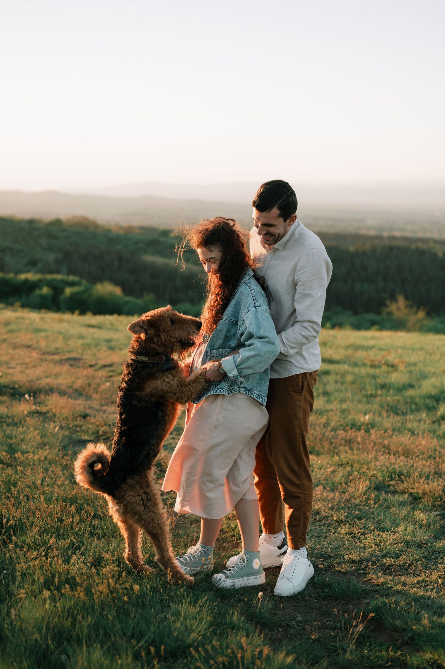 A couple and their fluffy dog play together in a grassy field during golden hour.