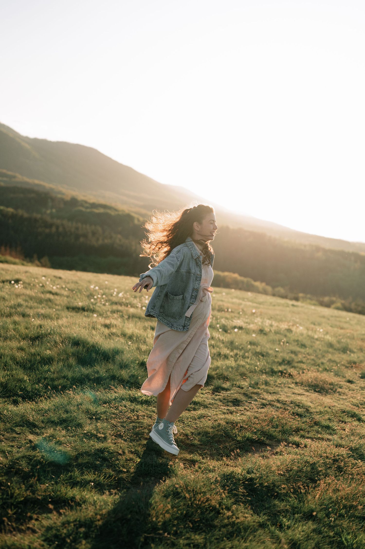 Person walking through a sunlit meadow on a mountainside during golden hour, creating a dreamy silhouette.