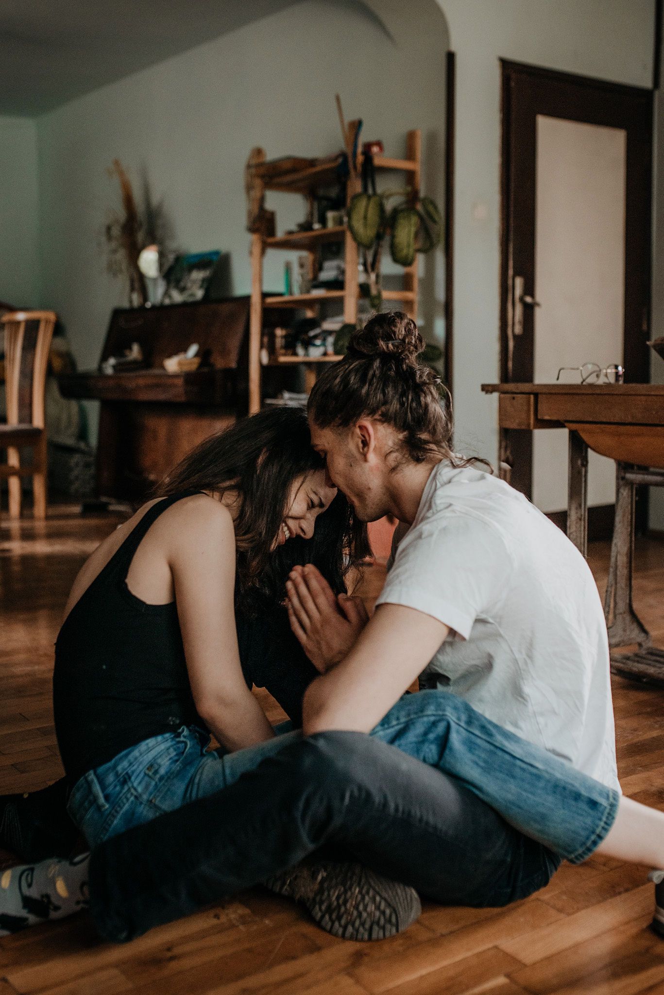 Two people in casual clothing lie close together on a hardwood floor in a domestic setting.