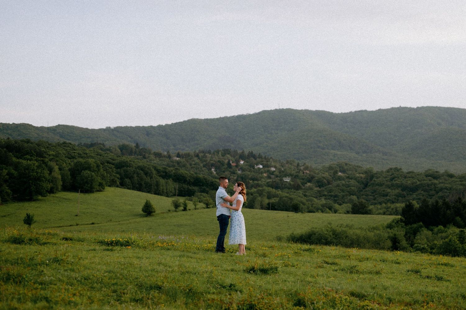 Mountain meadow with white tent structure and distant figures against forested hills.
