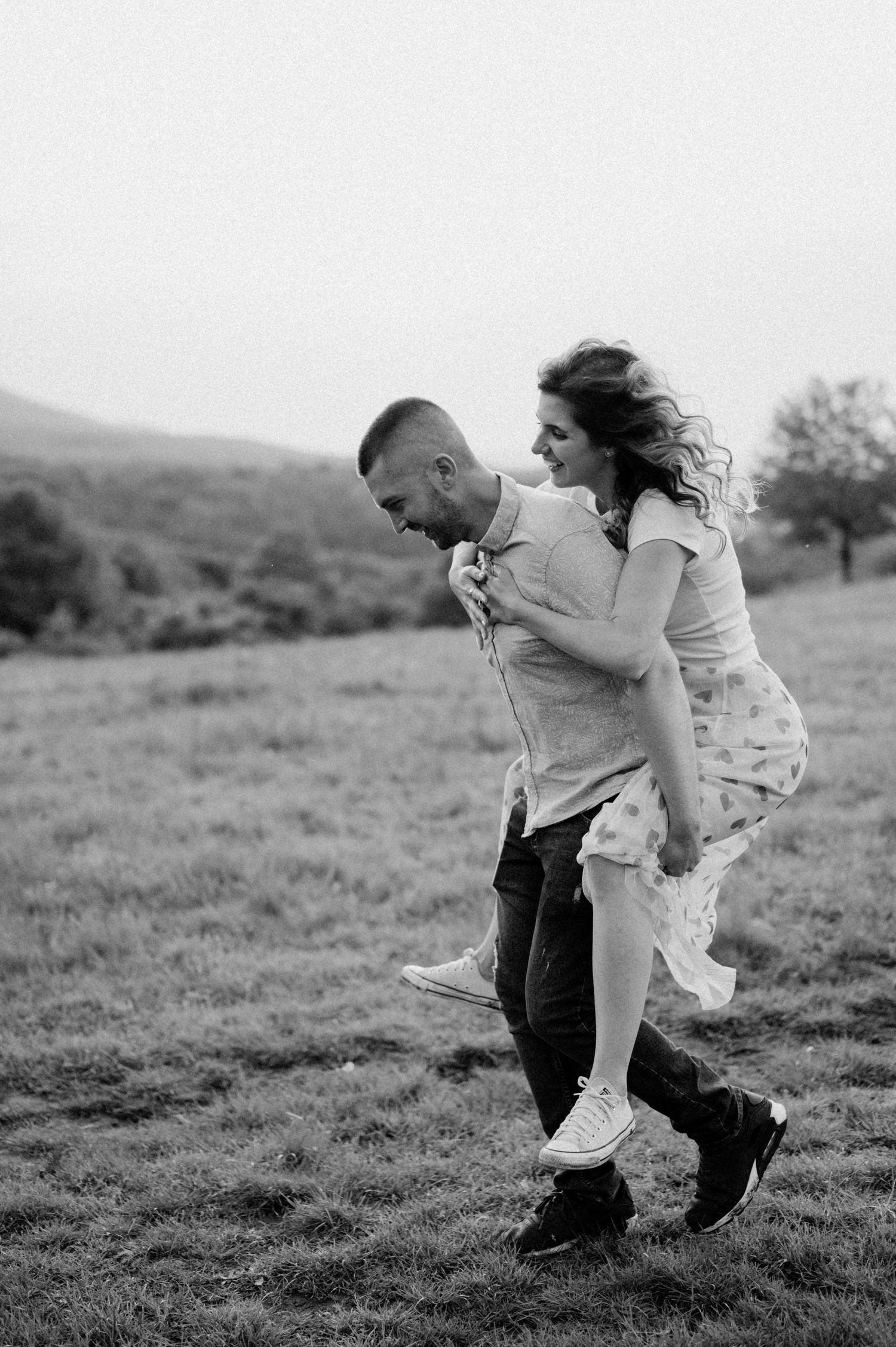 A black and white photo captures a couple playing and laughing in a meadow.
