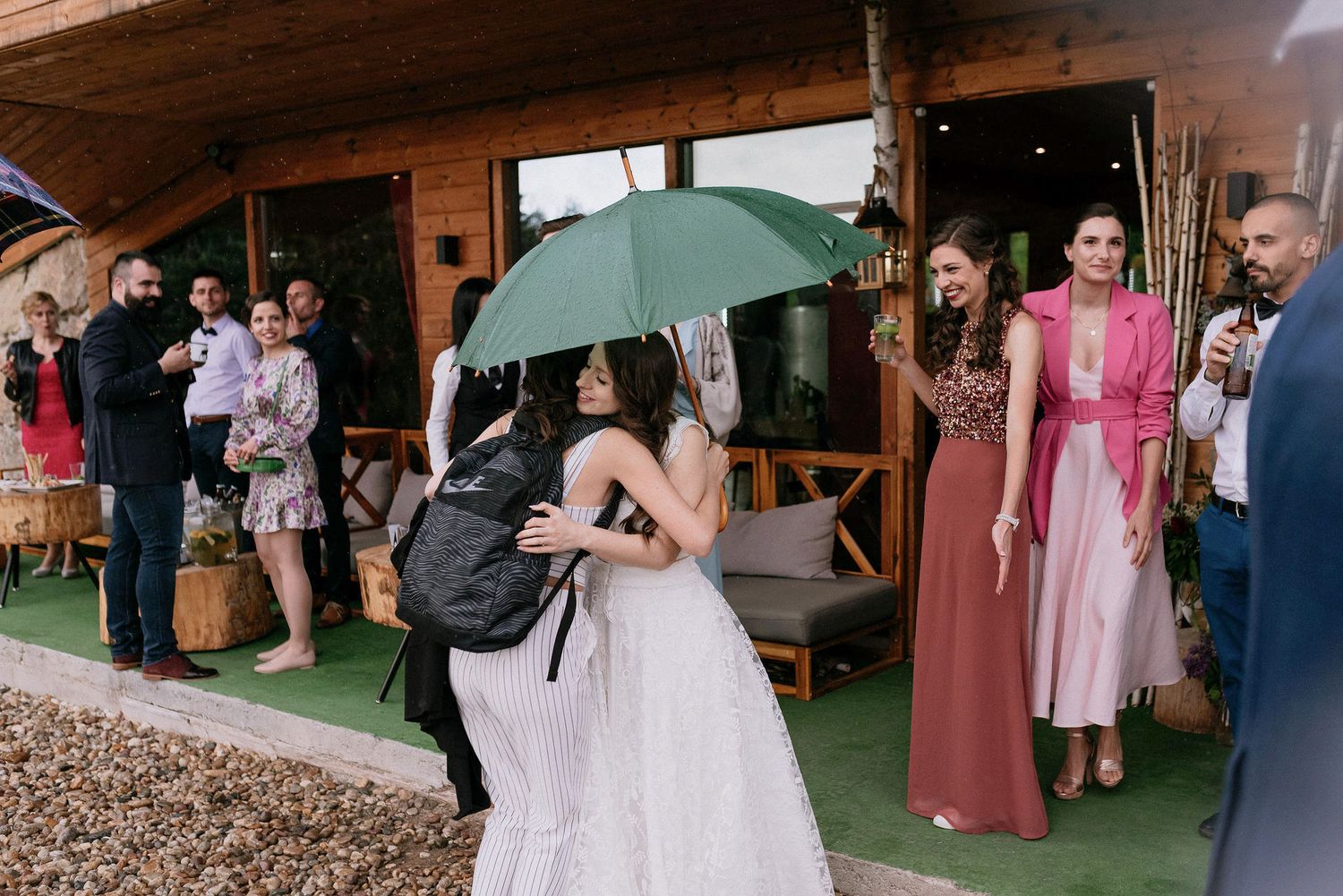 Wedding guests gather under umbrellas outside a rustic wooden venue during light rain.
