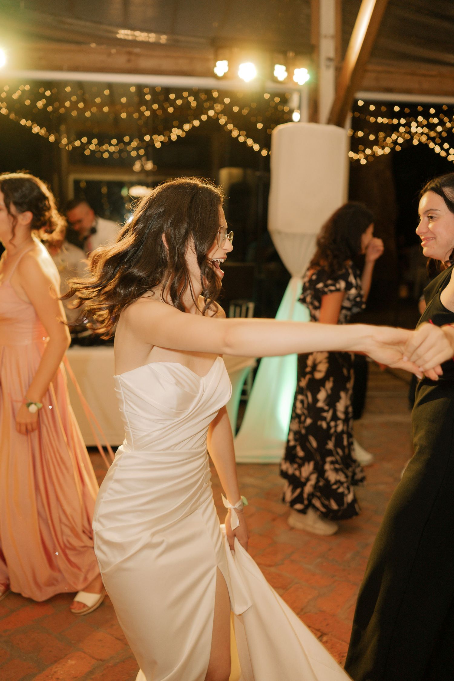 Wedding party members dancing energetically on a brick floor under string lights at an indoor reception venue.
