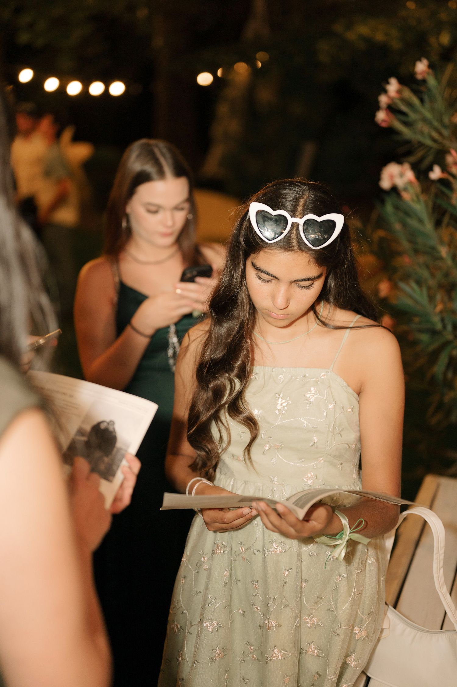 People reading at an evening event with string lights overhead and heart-shaped sunglasses on display.