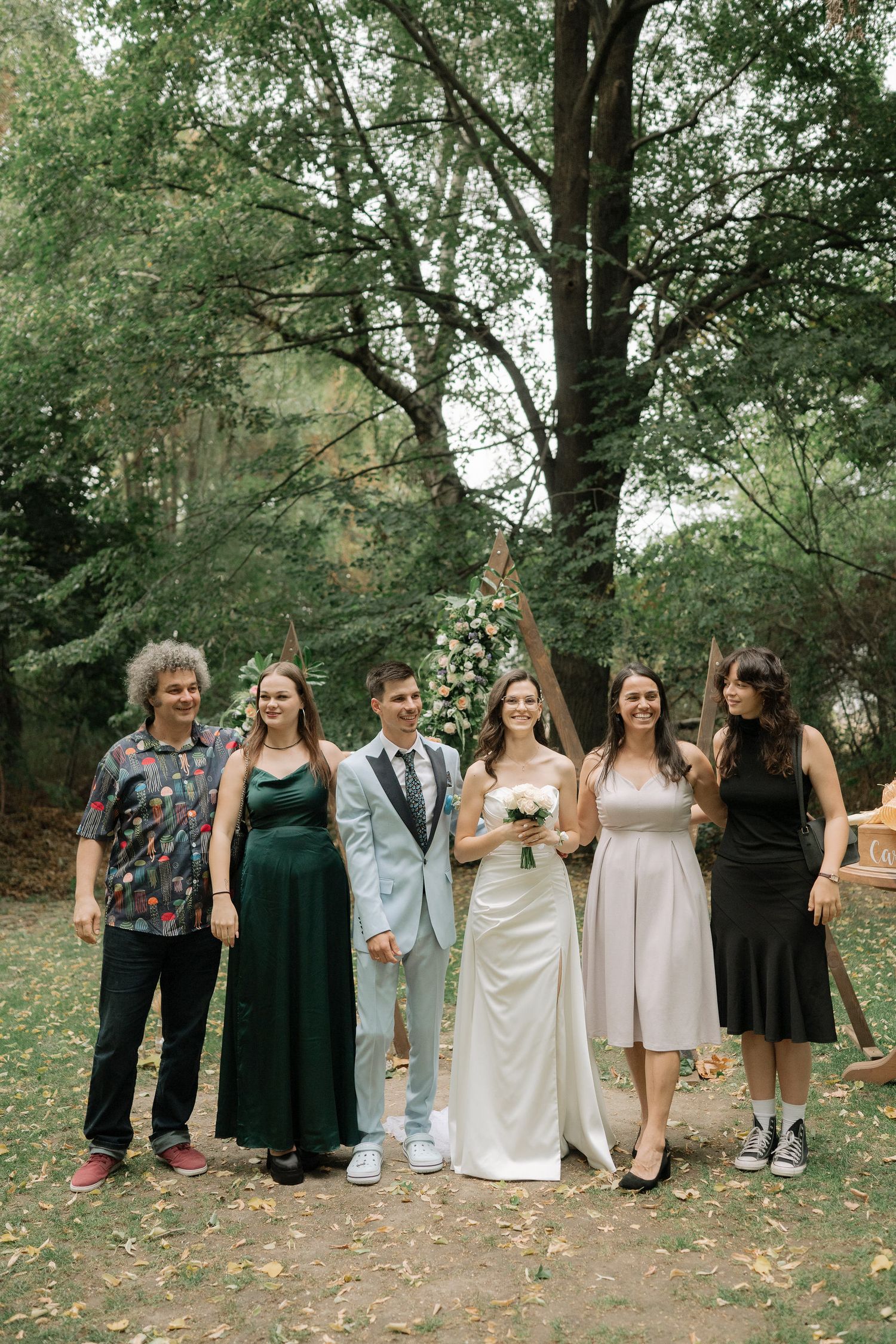 A wedding party poses outdoors in formal attire with bridesmaids in dark dresses and light dresses standing in a line.