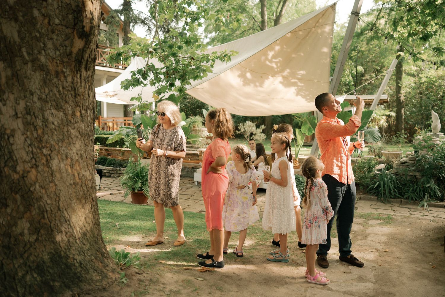 A series of outdoor wedding photos showing guests mingling under trees with parasols and summer attire at a garden venue.