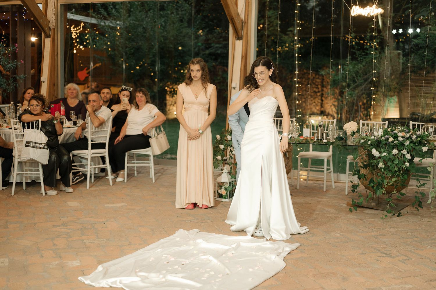 A wedding reception in a glass-enclosed venue with elegant decor, white chairs and greenery surrounding the space.