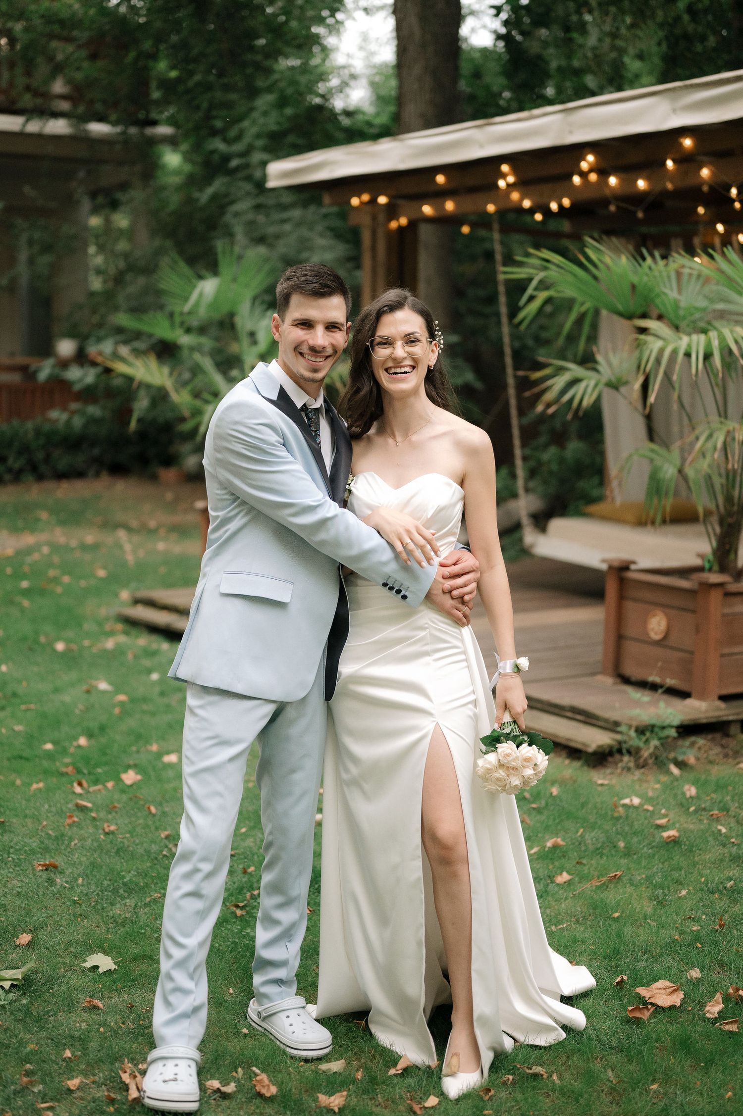 A couple shares a tender moment on their wedding day in a garden setting with string lights and greenery.