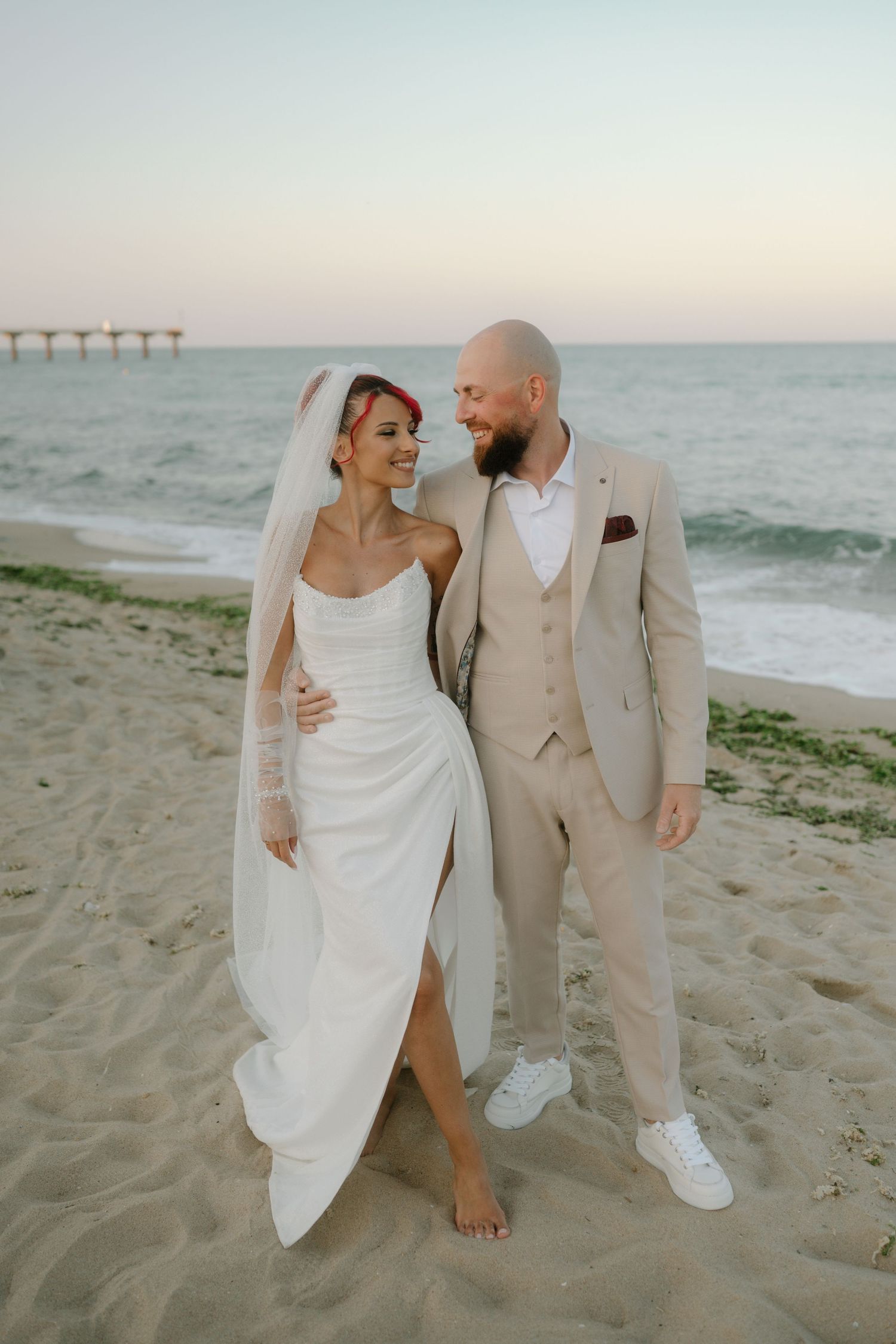 A romantic beach wedding scene at sunset with a couple posing on the sand, with a pier visible in the background.