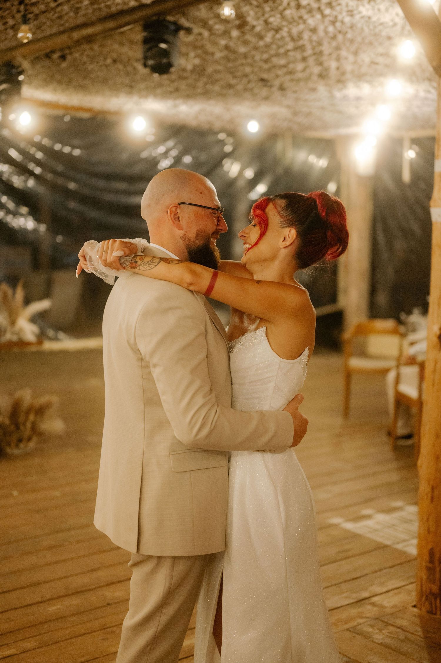 A couple shares an intimate dance moment on a rustic wooden floor under warm string lights and exposed brick walls.