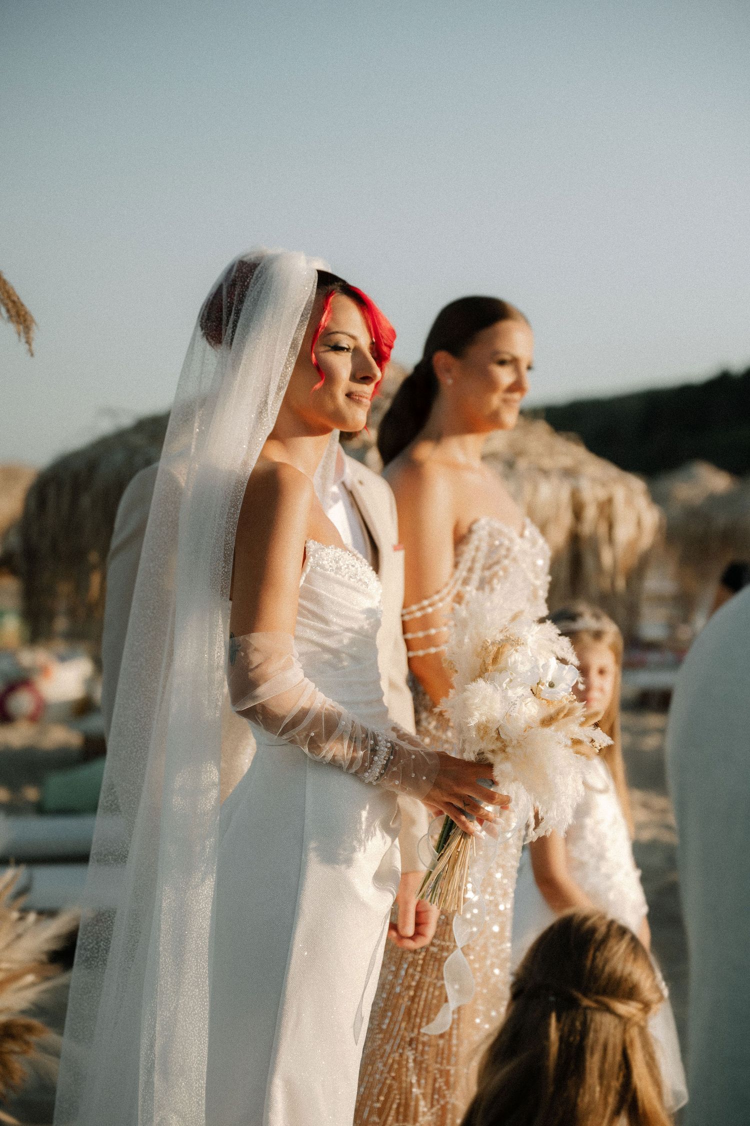 Three brides in elegant white and gold dresses stand together on a balcony during sunset.