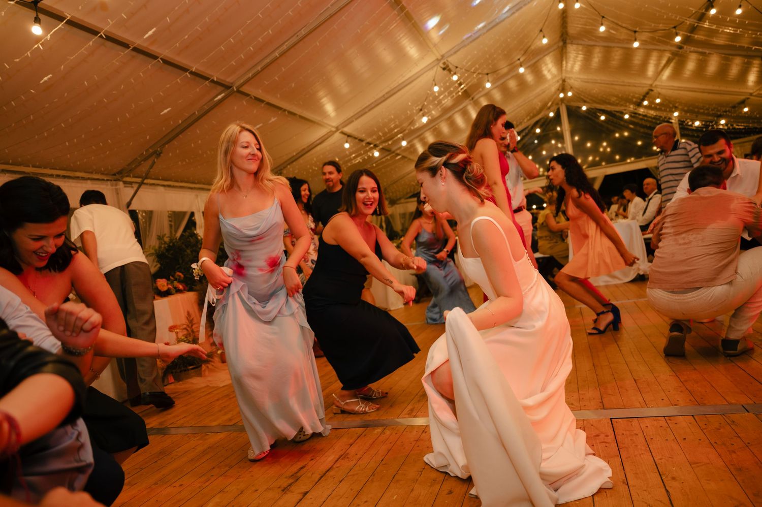 People dancing and celebrating at a wedding reception on a wooden dance floor under warm lighting and decorations.