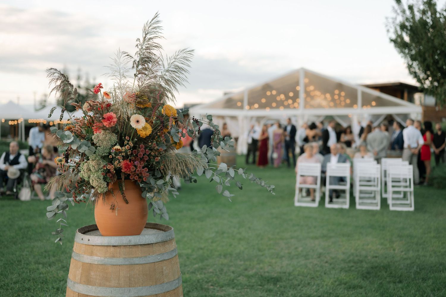 Rustic wedding reception setup outdoors with floral arrangement on wine barrel and white chairs under tent at sunset.