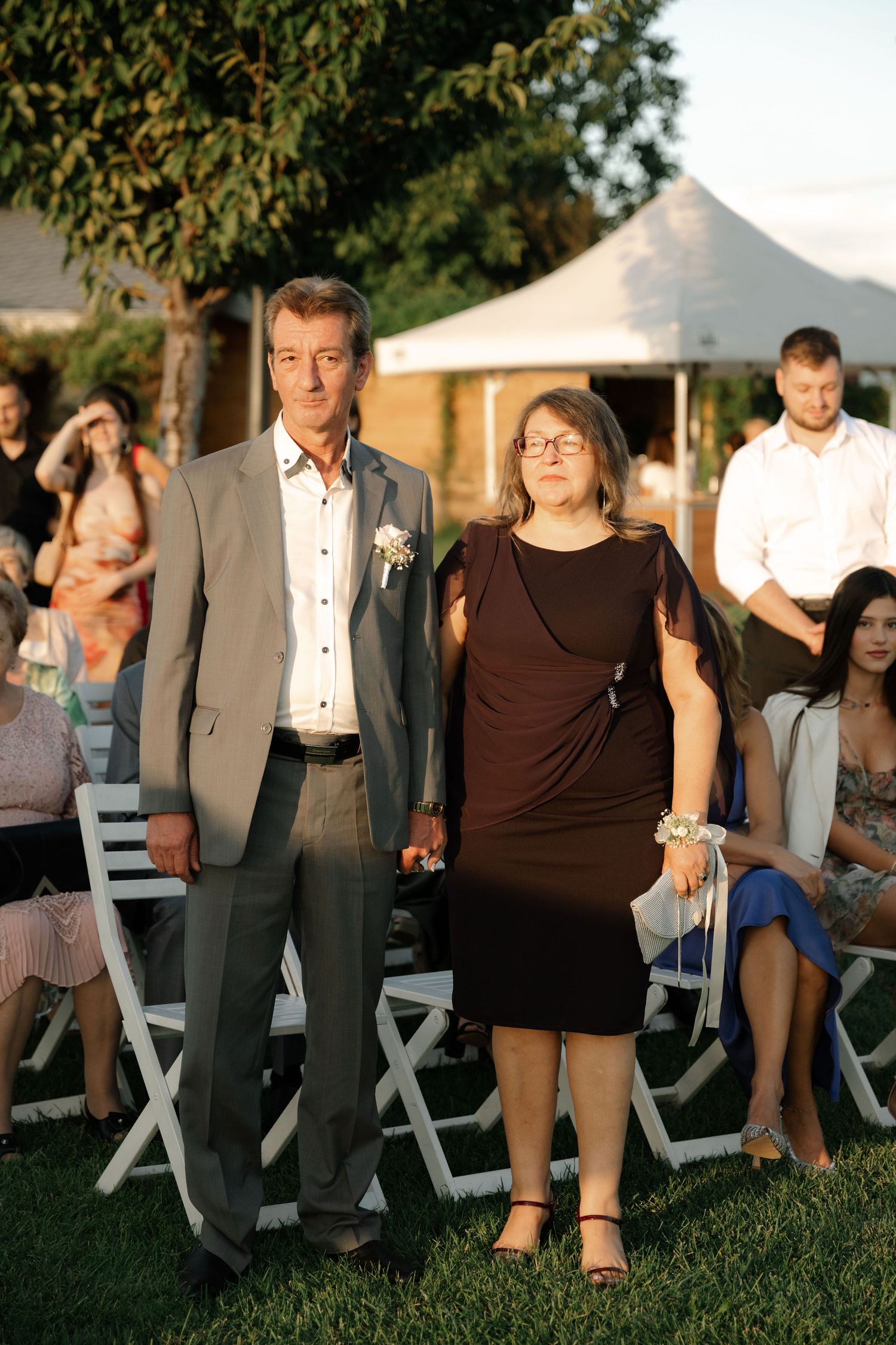Wedding guests dance and celebrate on a grassy lawn with white tents in the background during sunset.