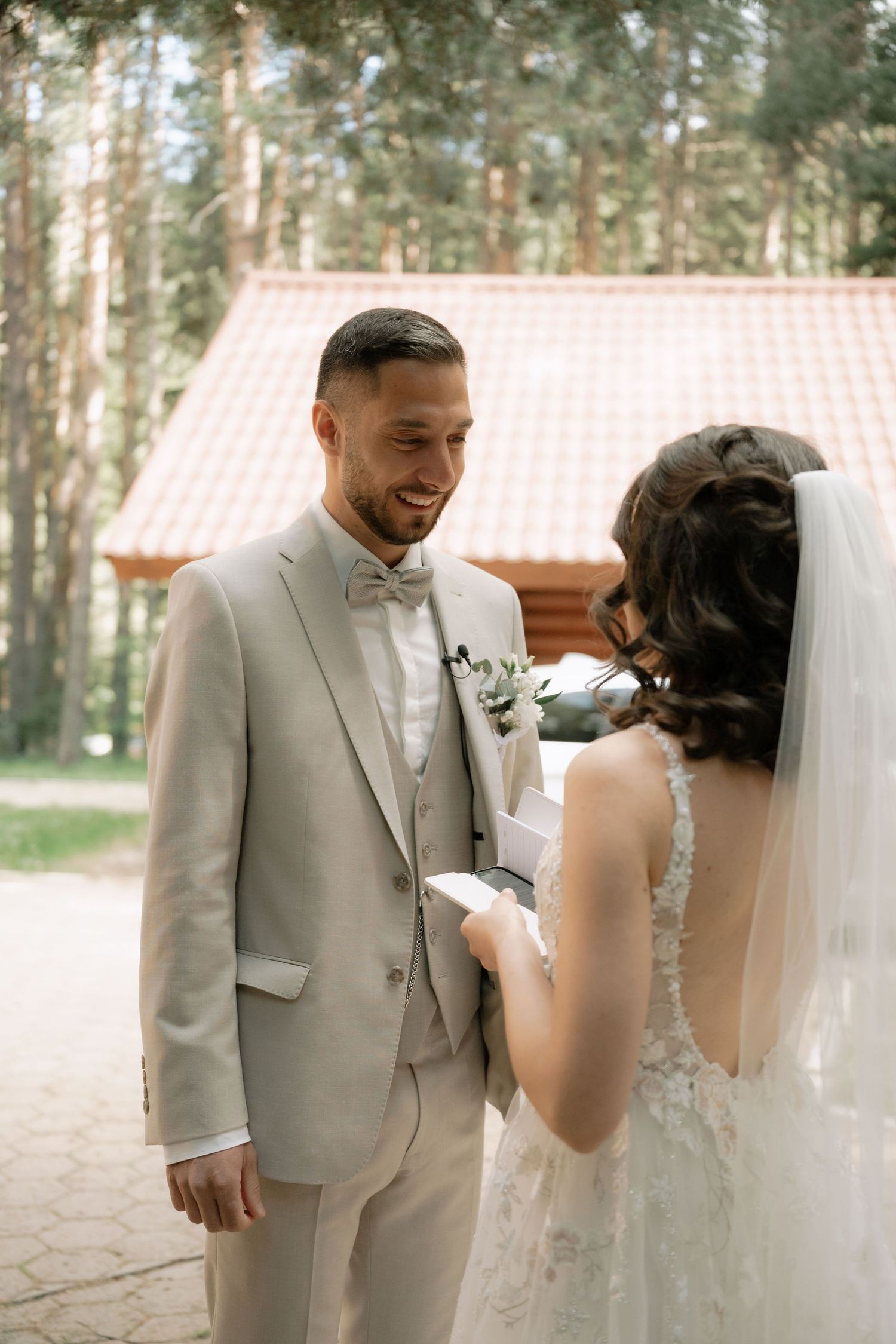 Smiling groom in beige suit reads wedding vows with bride in lace dress and veil outdoors in forest setting.