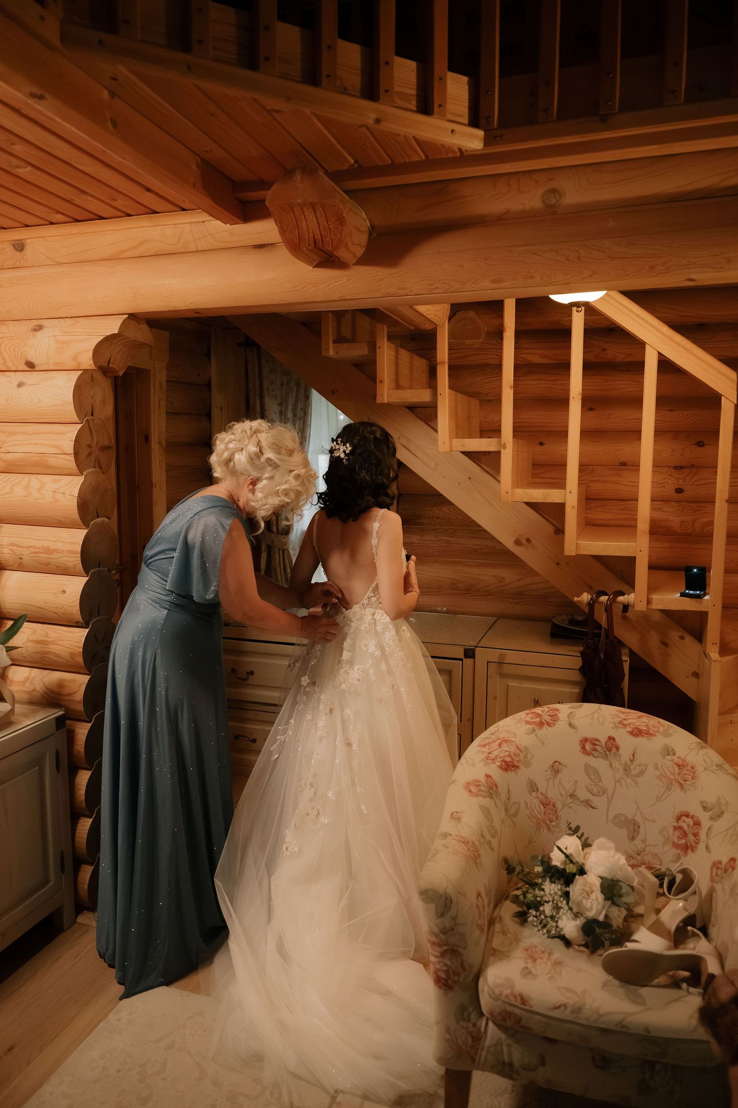 Bride in white gown being helped by woman in blue dress inside rustic log cabin with wooden staircase.