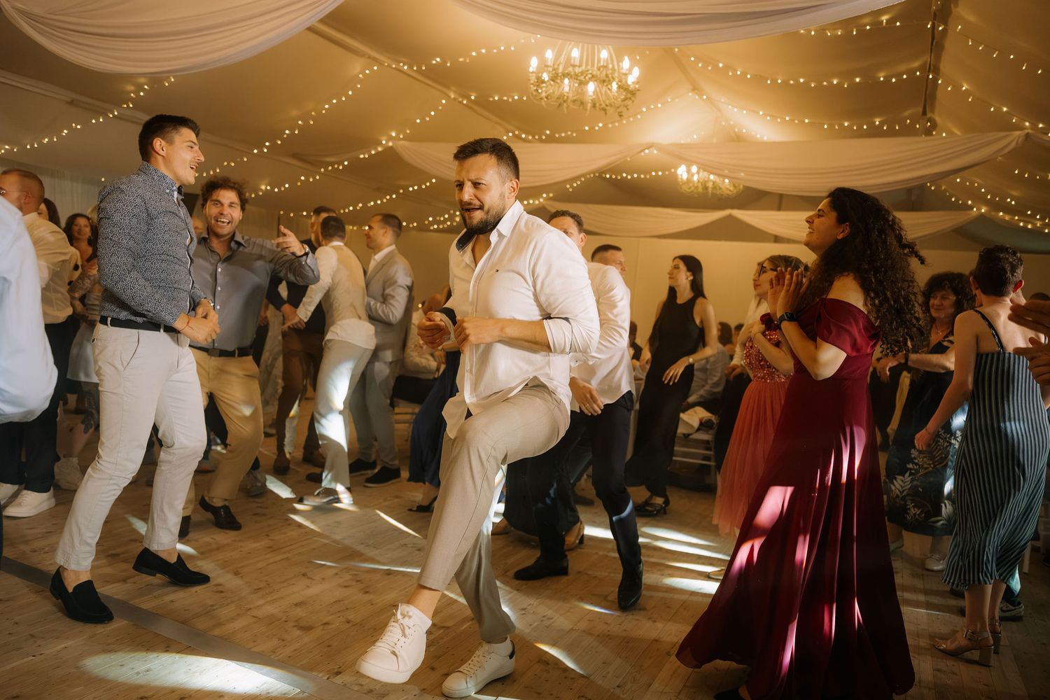 Guests dancing energetically at a wedding reception in a warmly lit venue with draped ceiling decor.
