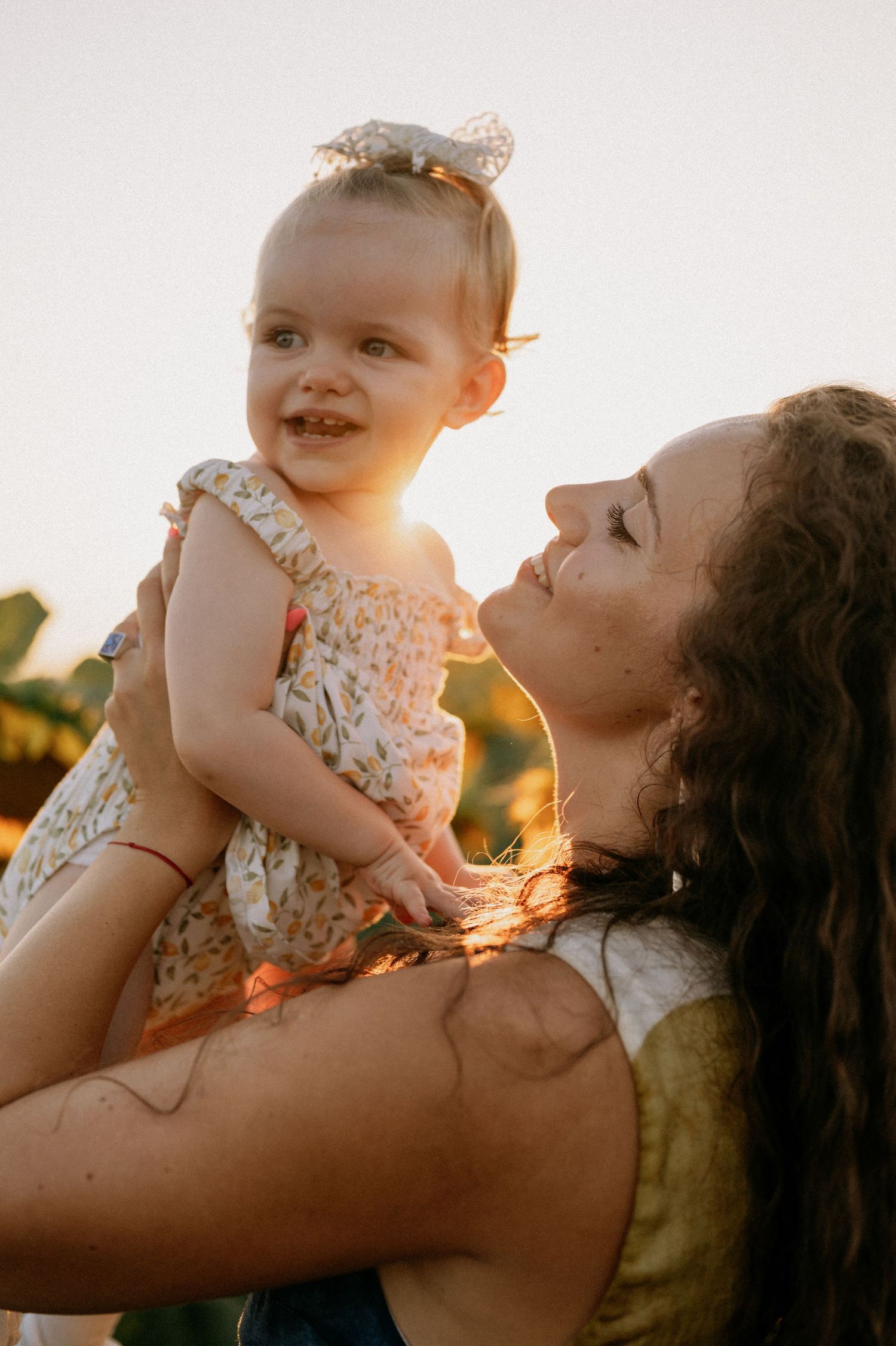 A mother lifts her smiling baby girl in a floral dress during a golden sunset outdoor photo session.