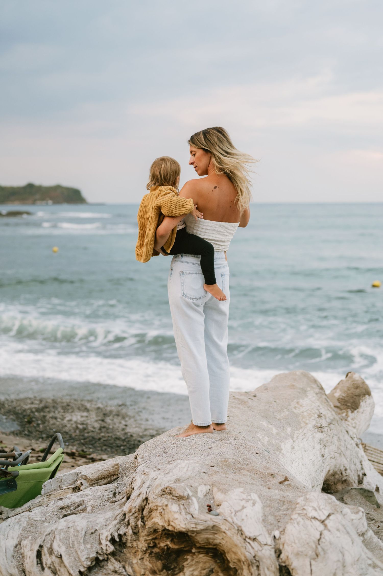 A woman holds a child while standing on driftwood at a beach, wearing white jeans and an off-shoulder top.