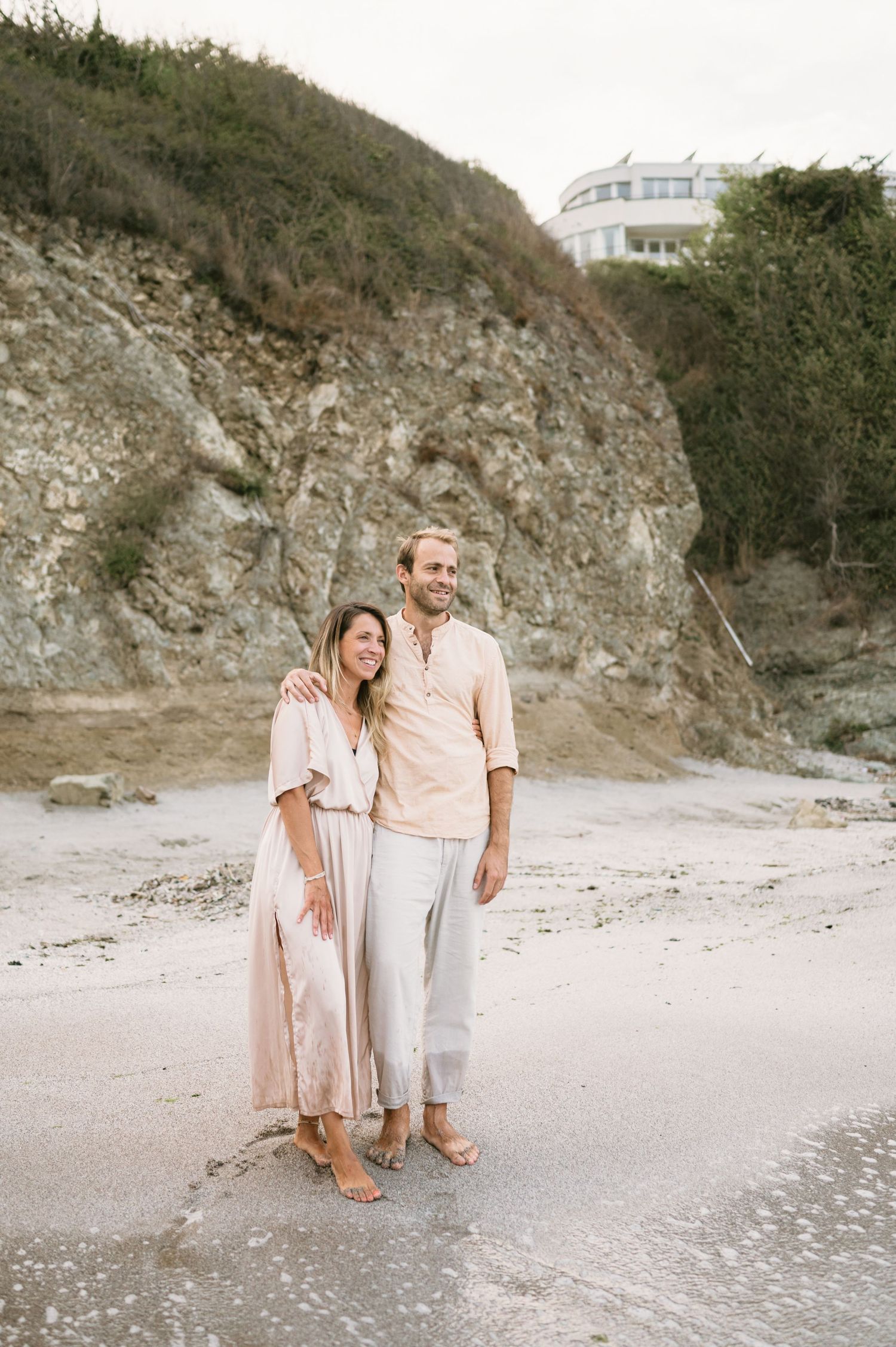 Couple in neutral tones embracing on a sandy beach with rocky cliffs and a building in the background.