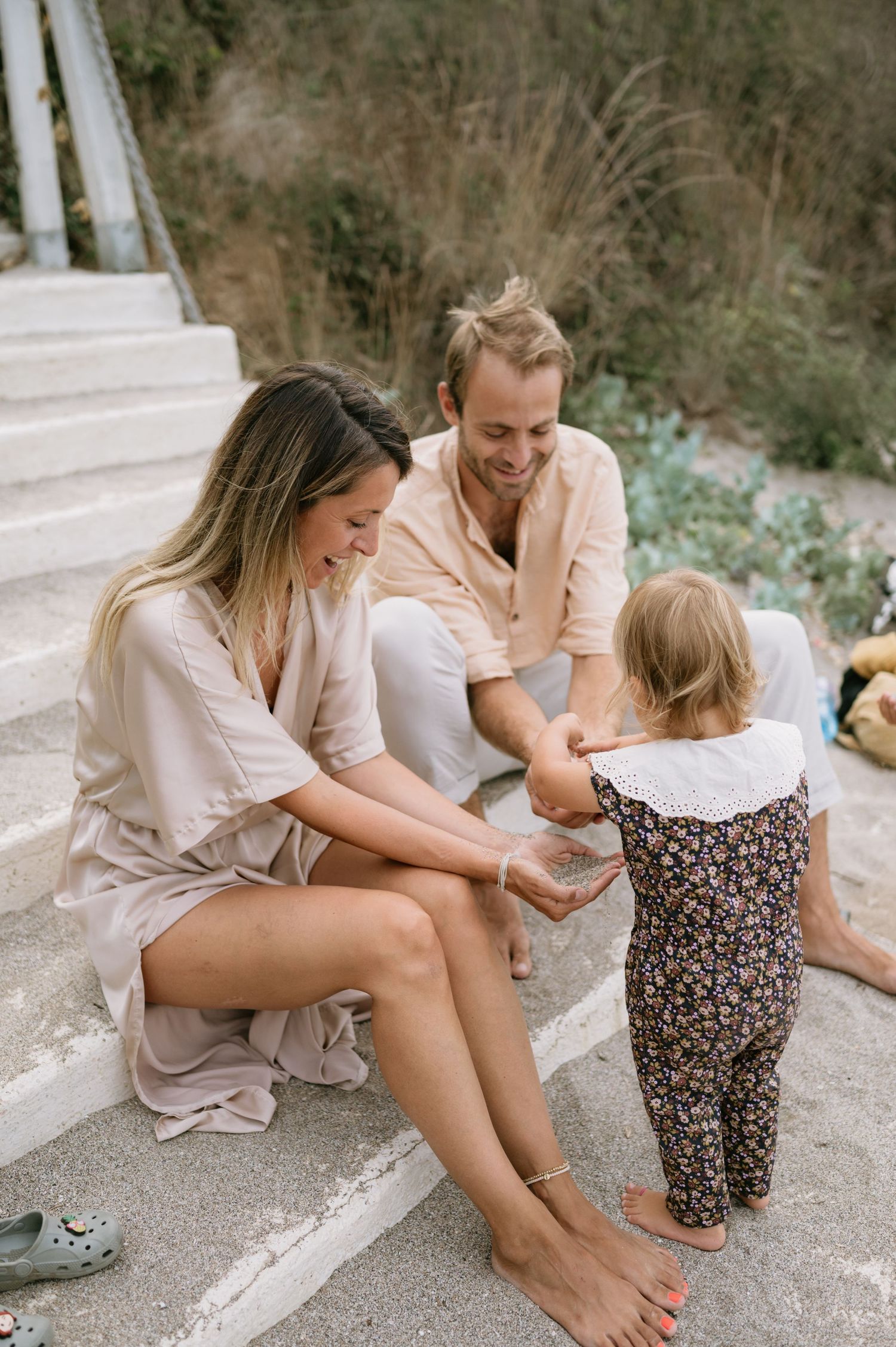 A happy young family sits on outdoor steps, smiling at their toddler daughter in a floral outfit.