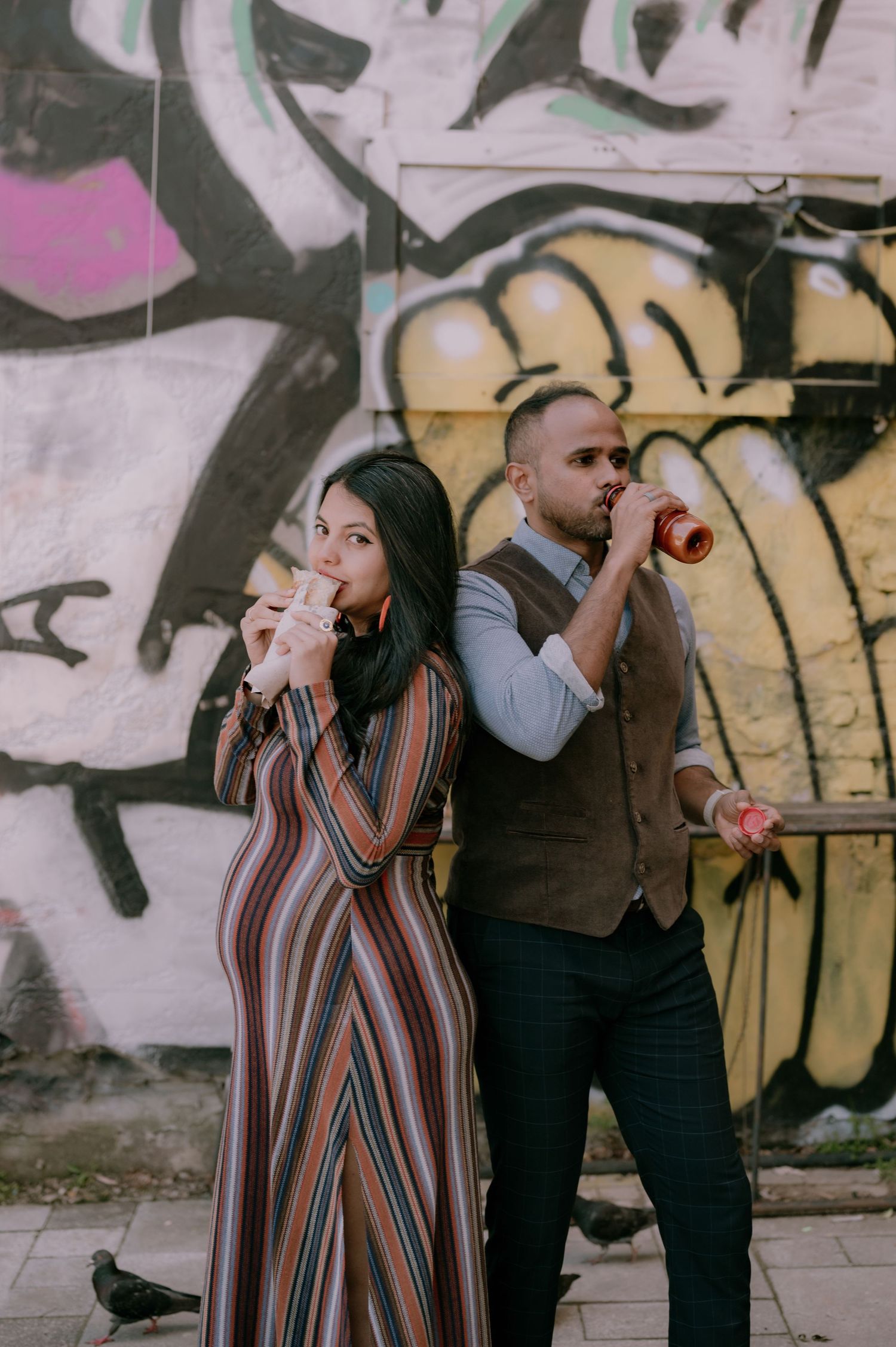 A couple poses back-to-back drinking from bottles in front of a colorful graffiti wall outdoors.