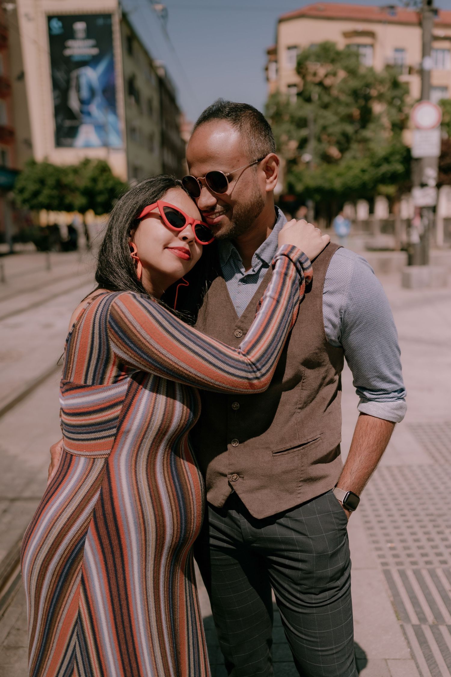 A stylish couple embraces on a sunny street, woman in striped dress with red sunglasses, man in vest and shades.