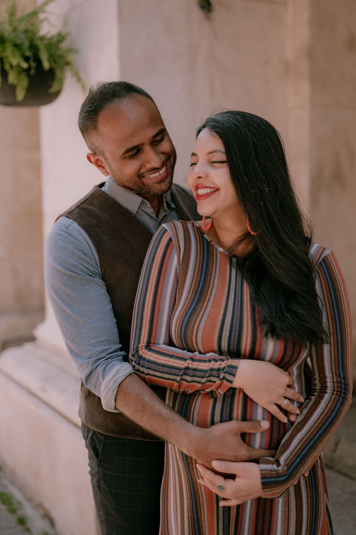 Smiling couple embracing outdoors during a maternity photoshoot, woman in striped dress cradling her baby bump.