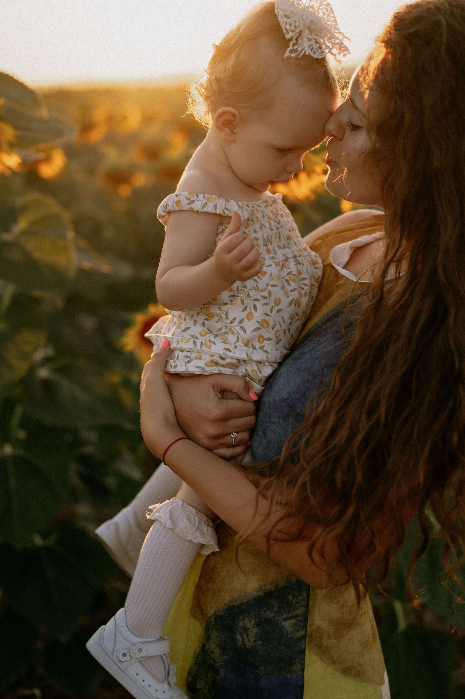 Mother holding baby girl in sunflower field at golden hour sunset, sharing a tender nose-to-nose moment.