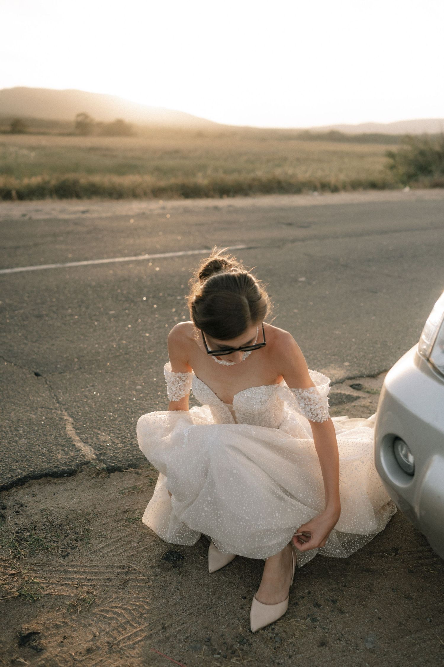 Bride in white off-shoulder lace gown adjusting her heel by a car on a rural road at golden hour sunset.